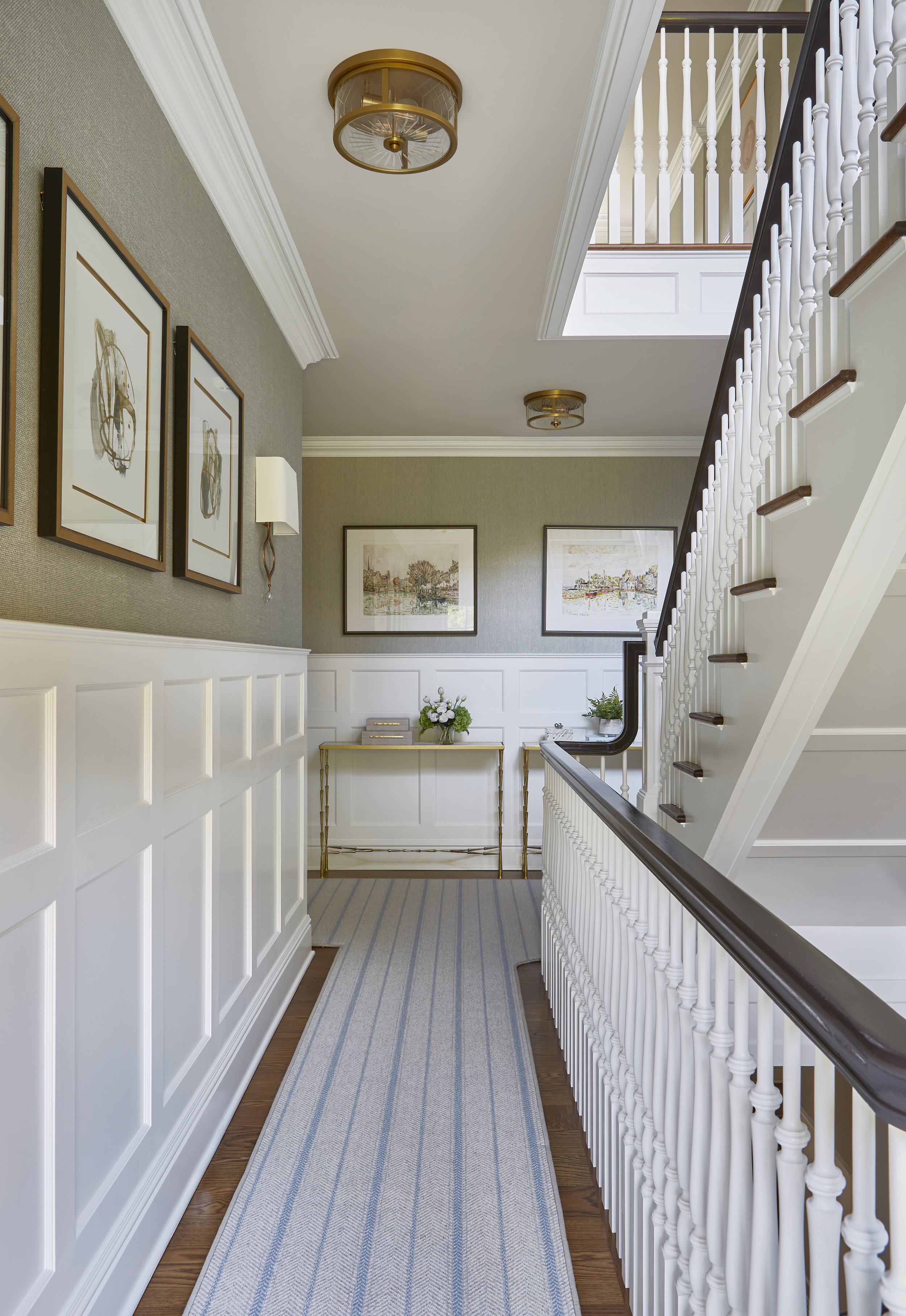Inside a well-lit hallway with white paneling, framed artwork, a striped rug, a gold console table with decorative items, and a staircase with white balusters and dark wood handrail.