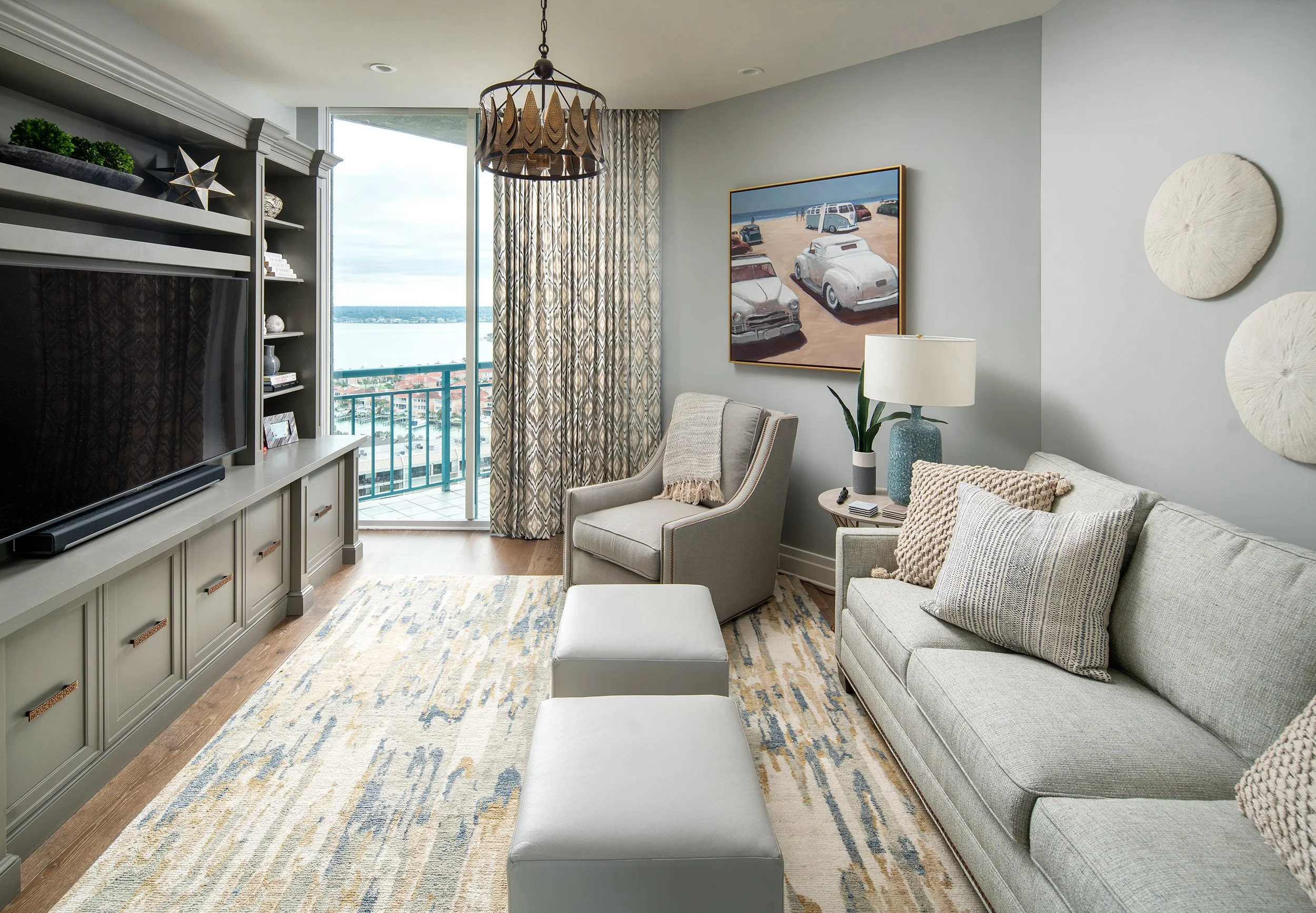 Living room with a large window and a balcony, light gray walls, a gray sofa with pillows, a beige armchair with a blanket, a side table with a lamp and plant, a patterned rug, a large television, and decorative plates and star sculptures, with a pai