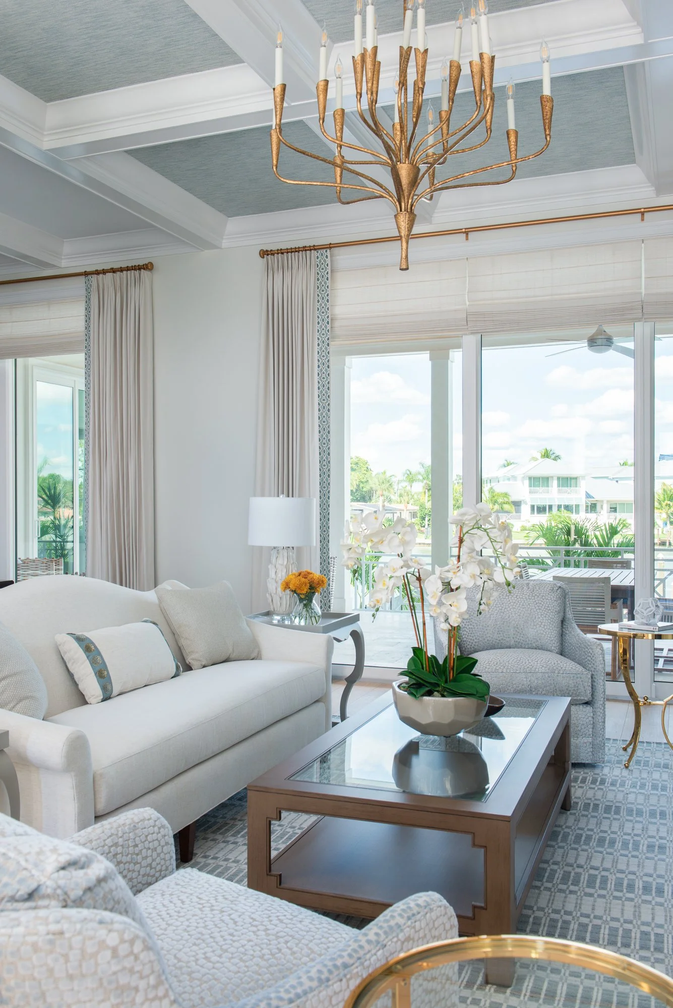 Living room with white sofa, armchair, coffee table, orchid plant, window with balcony view, chandelier, and decorative curtains.