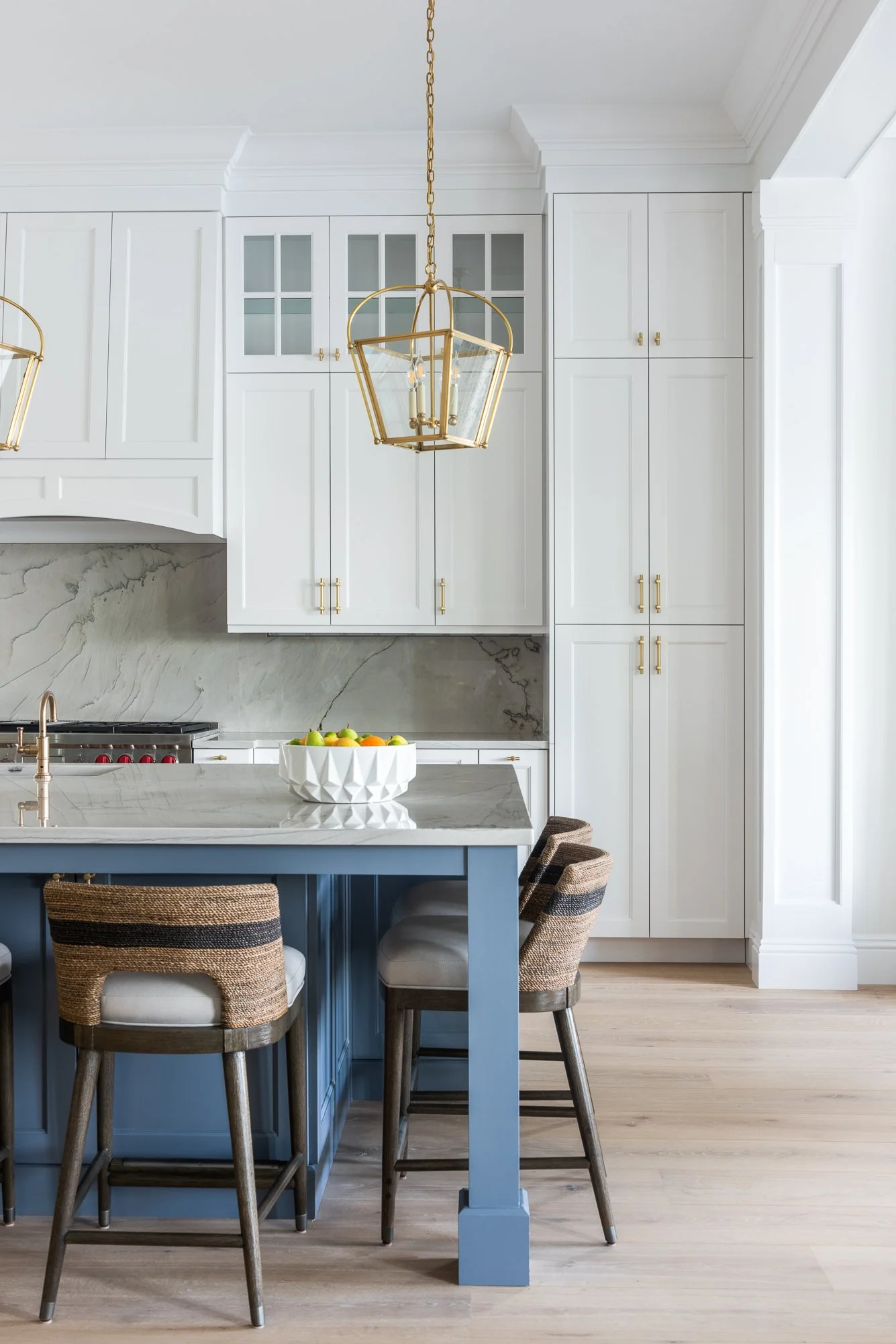 A modern kitchen with white cabinets, a marble backsplash, a blue kitchen island with bar stools, and gold pendant lighting.