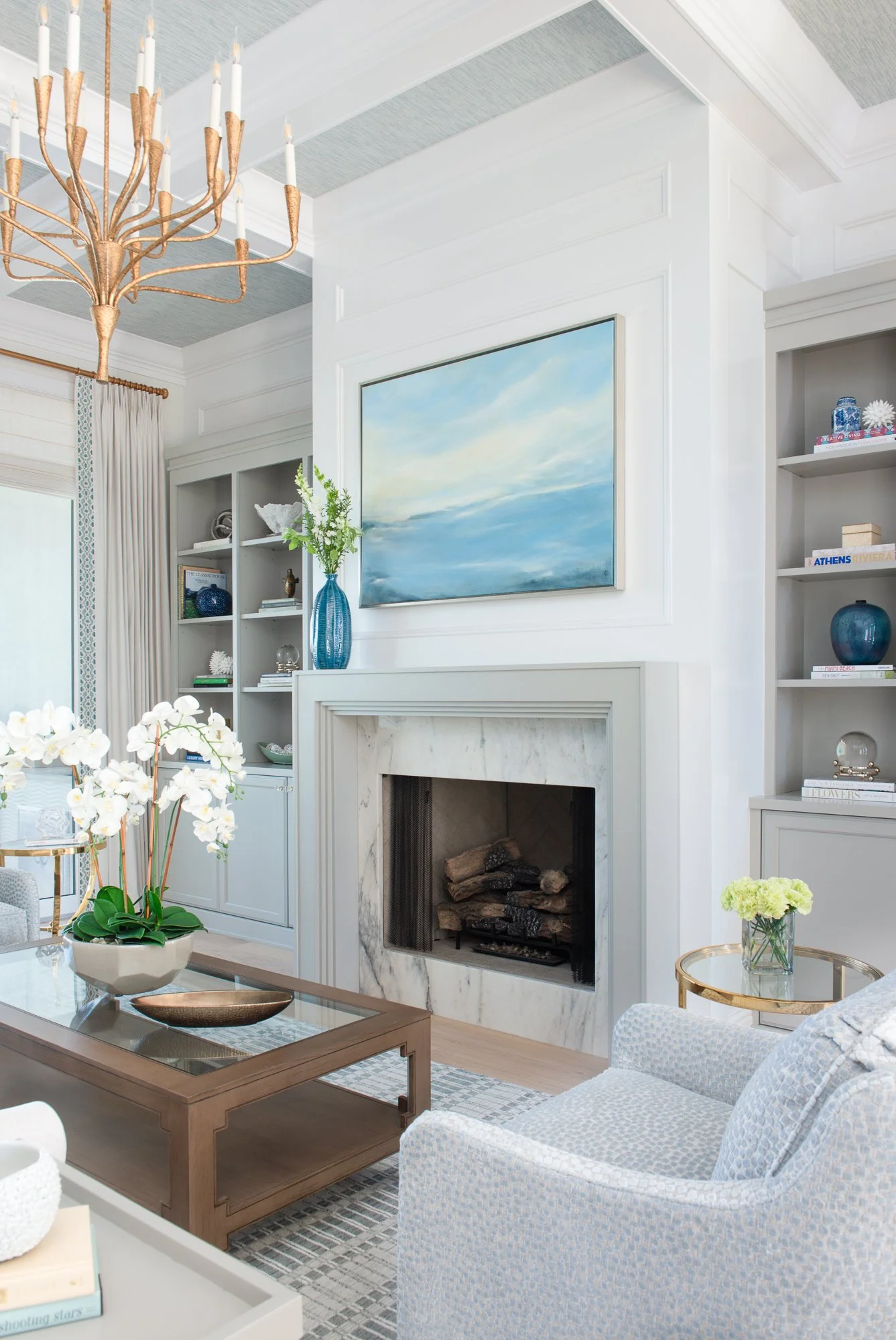 Living room with white walls, a marble fireplace, built-in bookshelves, a blue painting above the fireplace, a copper chandelier, a vase with flowers on the mantel, a white armchair, a wooden coffee table with a white orchid, and a side table with a 