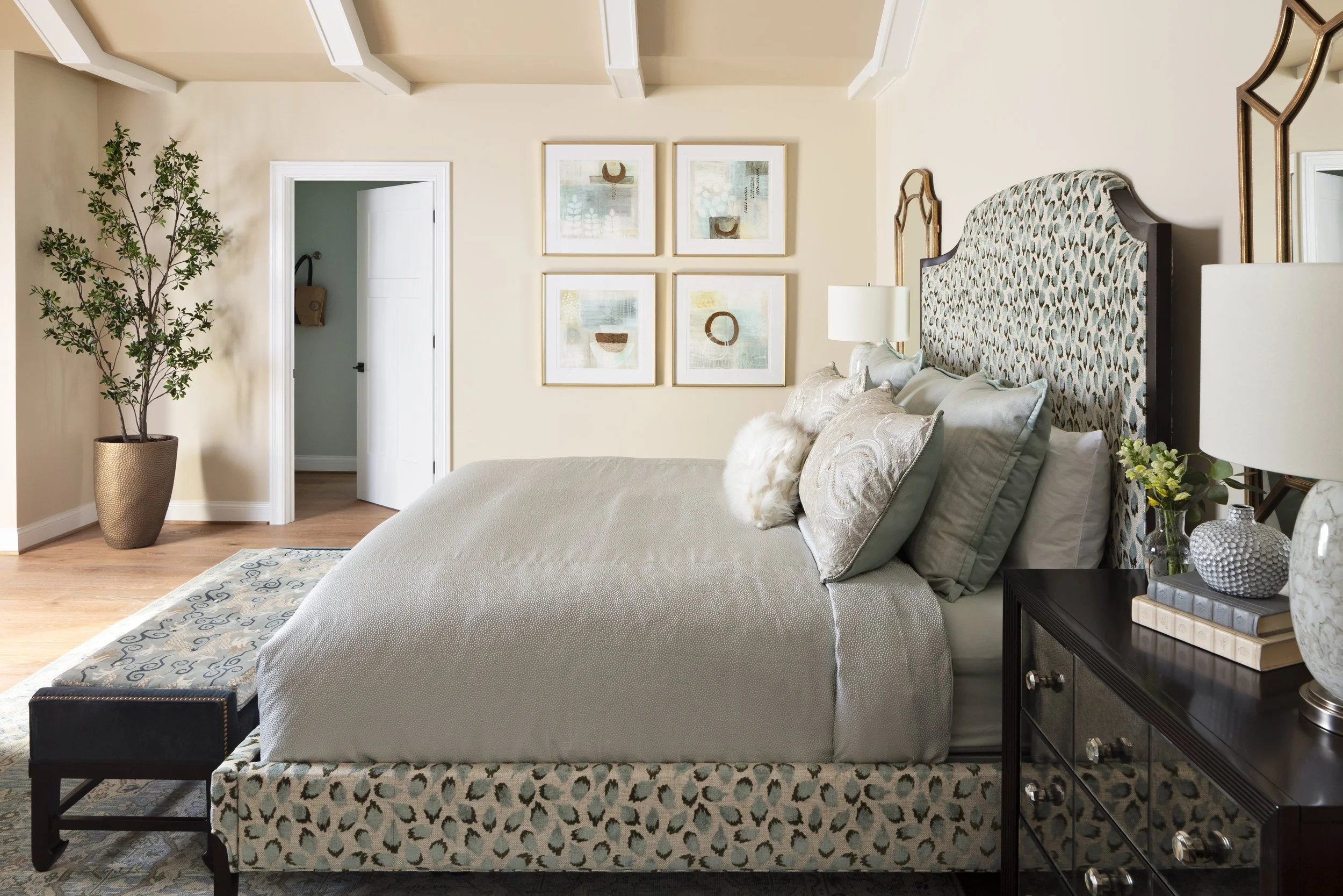 A neatly made bed with a patterned headboard, several decorative pillows, and a beige bedspread. Next to the bed is a black bedside table with a lamp, decorative vase, and books. The wall behind the bed has four framed abstract artworks. To the left,
