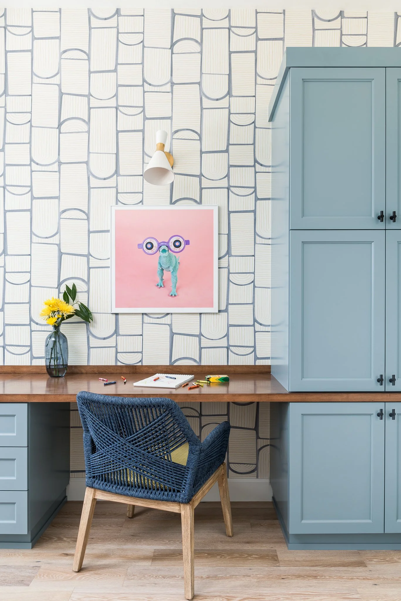 A home office with a desk, blue woven chair, blue cabinets, wallpaper with geometric pattern, framed picture of a frog with glasses, vase with yellow flowers, and some scattered crayons and paper on the desk.