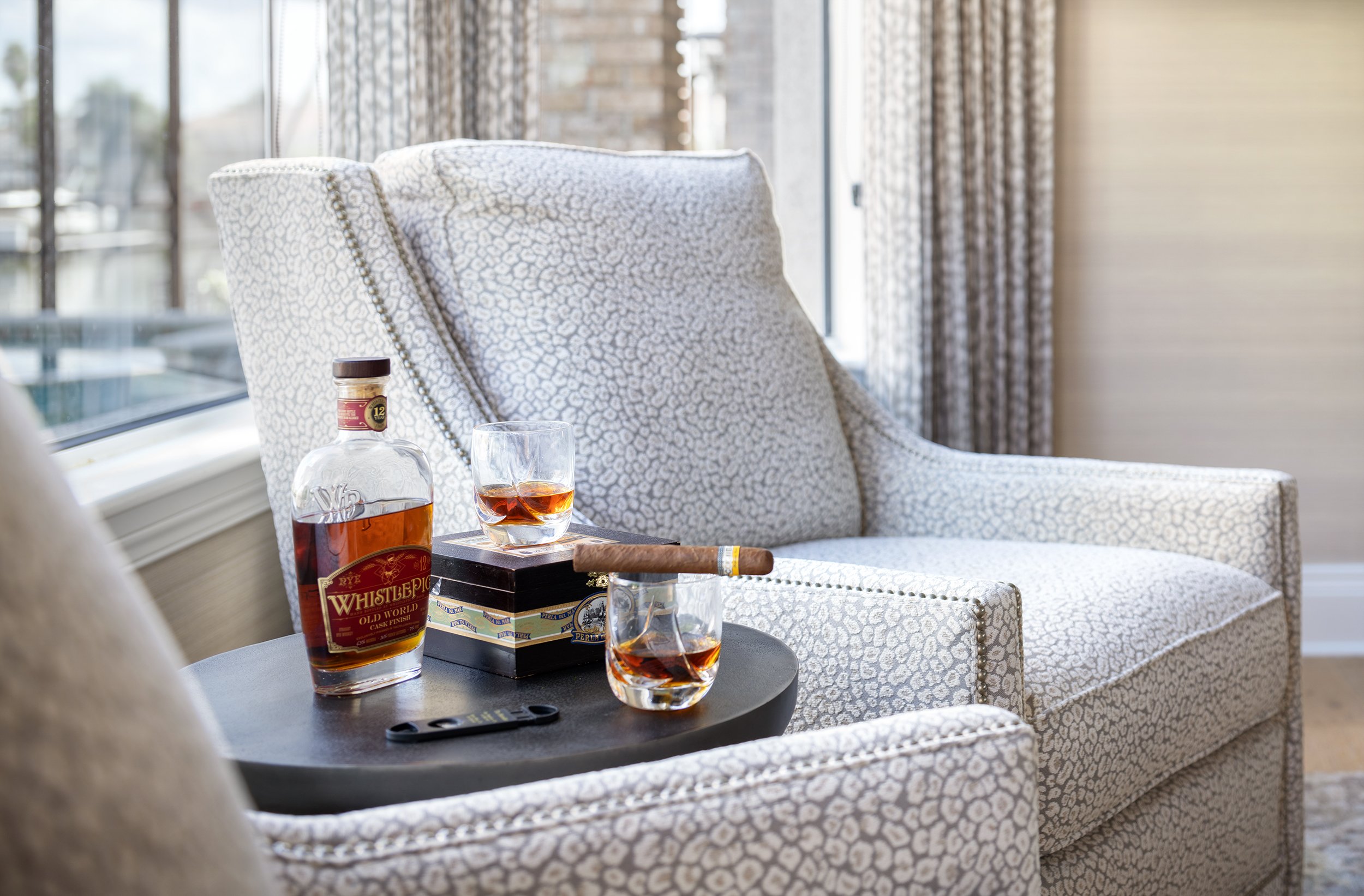 A living room with patterned armchair, a round black table holding a bottle of whiskey, two glasses with whiskey, a cigar, a cigar box, and a remote, near a window with curtains.