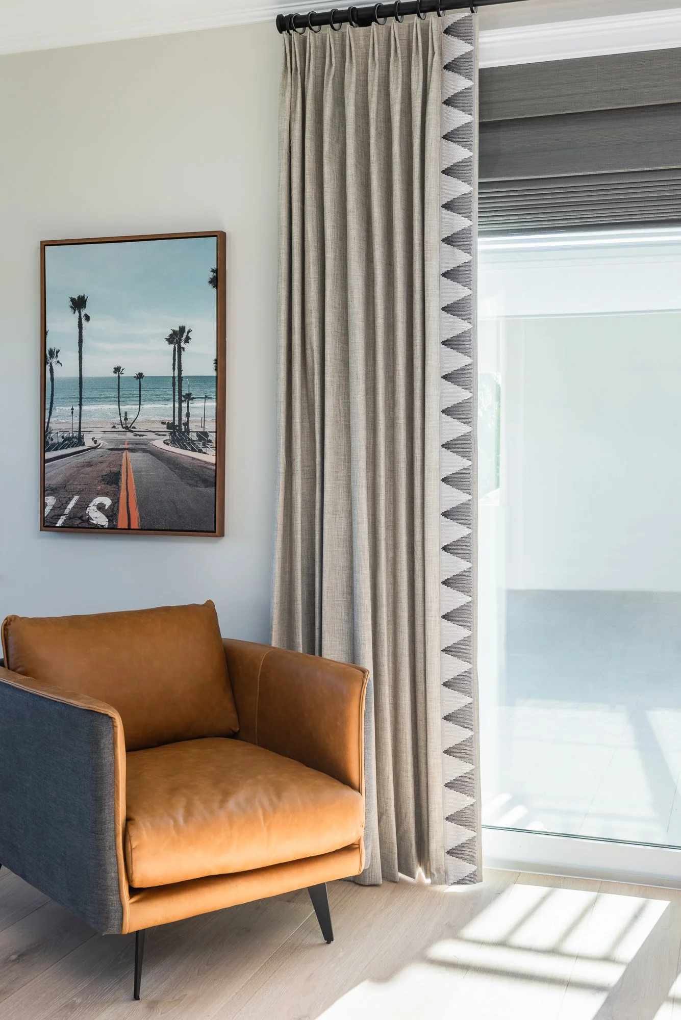 A living room with a brown leather armchair, a window with beige curtains and a view of a balcony, and a framed picture of a beach with palm trees on the wall.