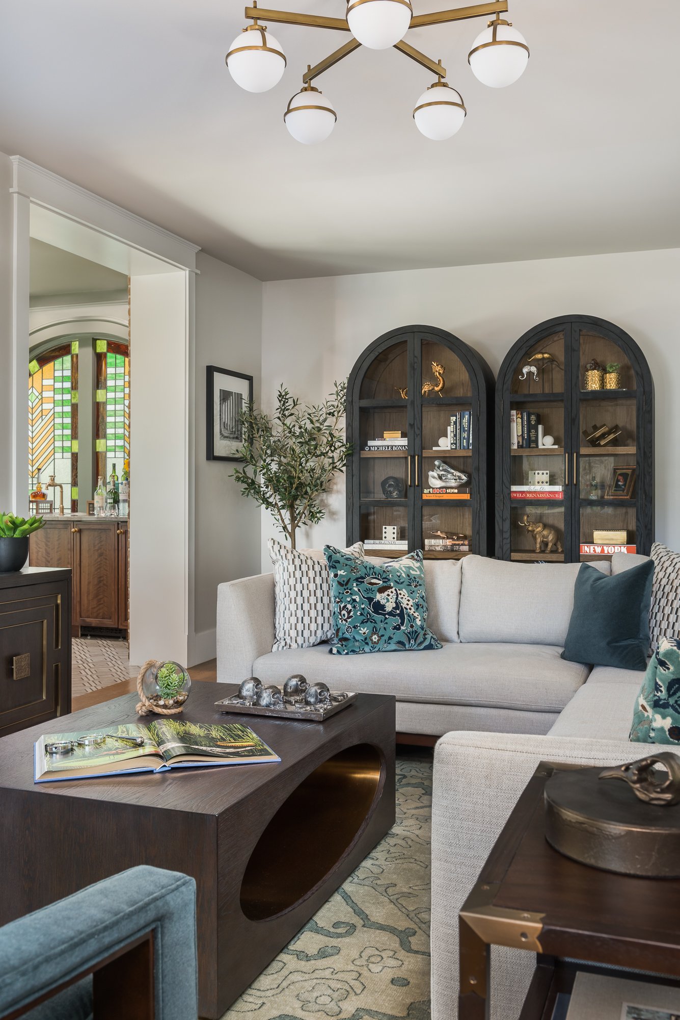 Living room with white sofa, decorative pillows, dark wooden coffee table with an open book, glass terrarium, and decorative metallic items. Black arched bookshelves with books and sculptures. Green potted plant, stained glass window in the adjacent room, modern chandelier with six white globes.