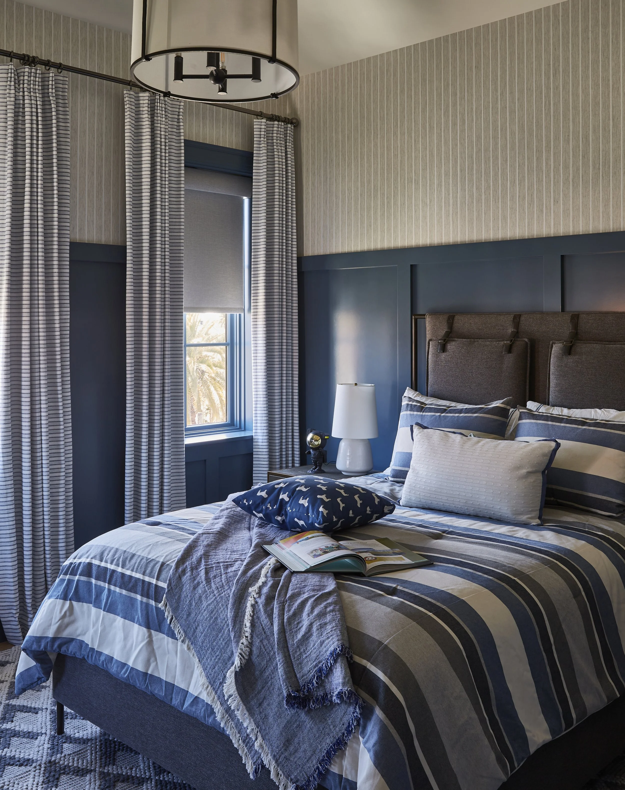 A neatly made bed with blue and white striped bedding, a gray throw blanket, and multiple pillows, one with a dog pattern. A window with striped curtains is on the left, with palm trees visible outside. A nightstand with a white lamp and a small cloc