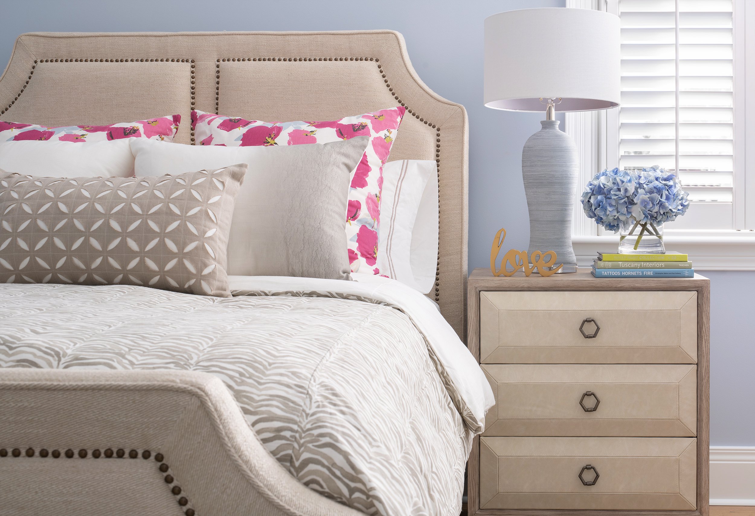 A bedroom with a beige upholstered bed, pink floral pillowcases, a beige decorative pillow, and a white comforter. A beige nightstand holds a large white lamp, a blue hydrangea arrangement, and a decorative "love" sign. White window blinds are in the