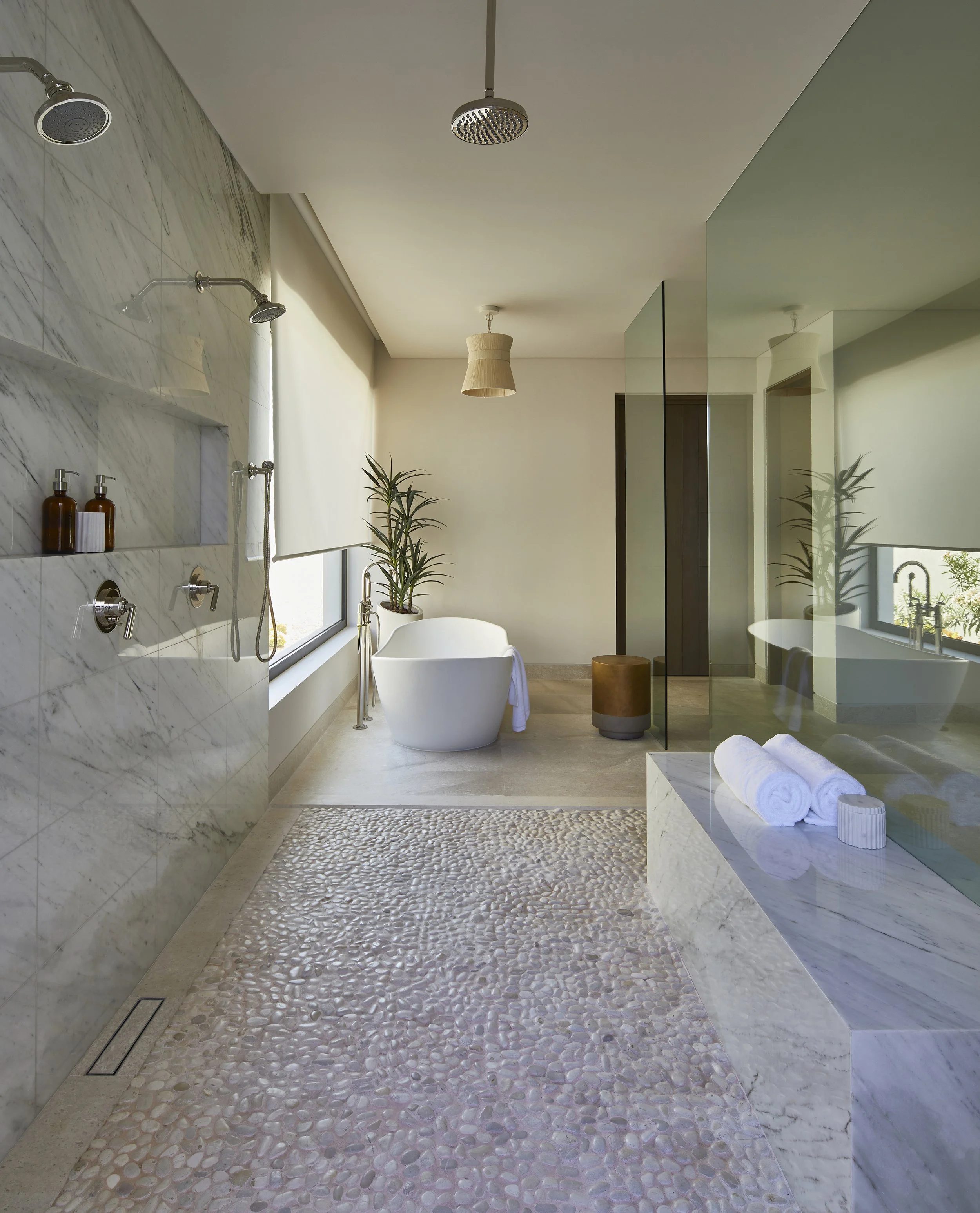 Modern bathroom with a freestanding bathtub, pebble floor, marble accents, shower area, potted plant, and large window with roller blind.