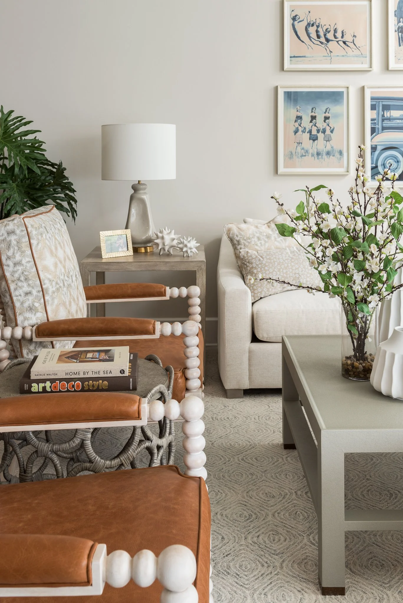 Living room with a beige sofa, patterned pillows, a gray coffee table with a large vase of white blossoms, framed artwork on the wall, a side table with a white lamp, a plant, and books on a chair.
