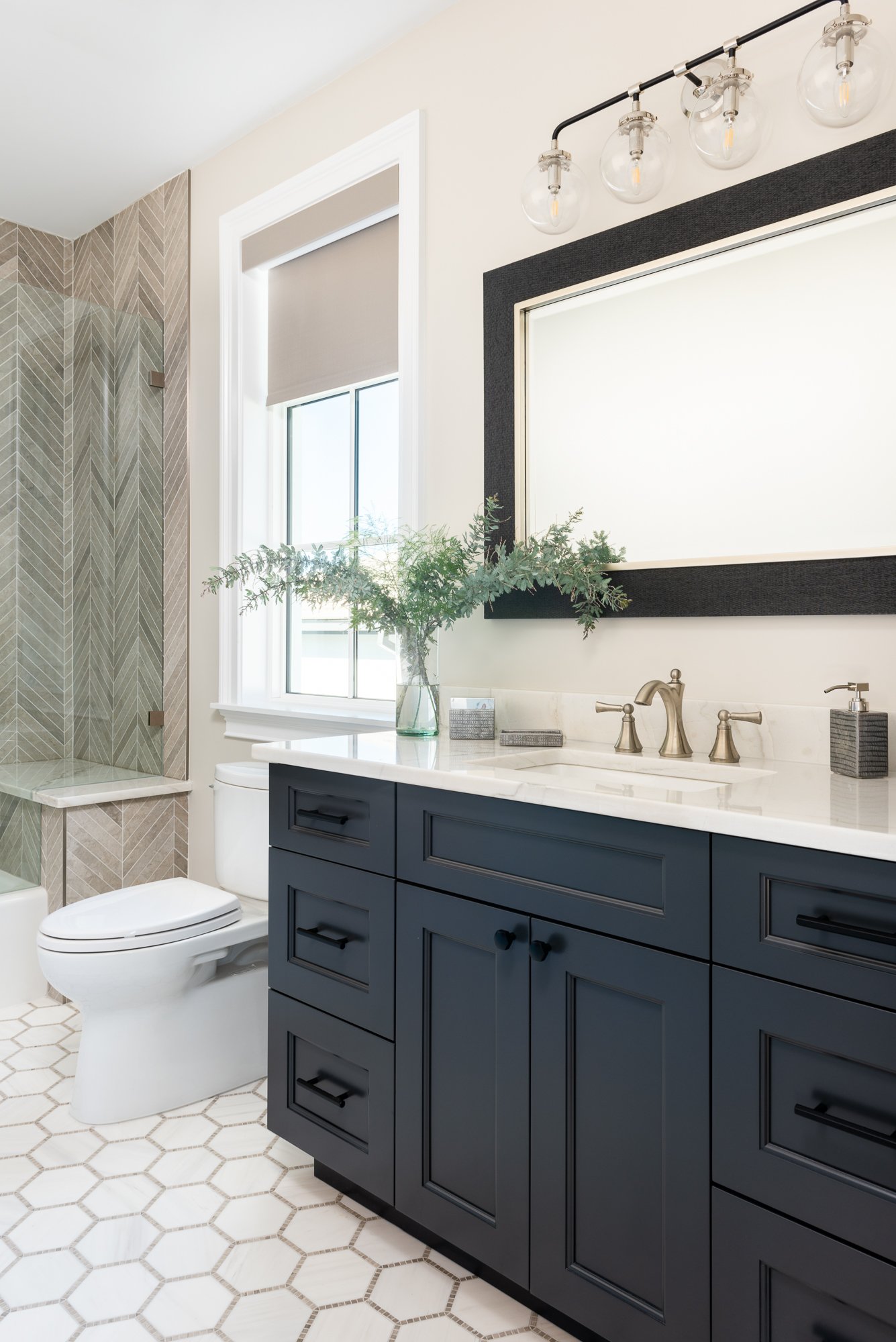 Modern bathroom with navy vanity, white marble countertop, large mirror, and hardware in brushed nickel or matte black; decor includes a vase of greenery and chrome fixtures; shower area with tan geometric tile pattern and bench; hexagonal tile floor