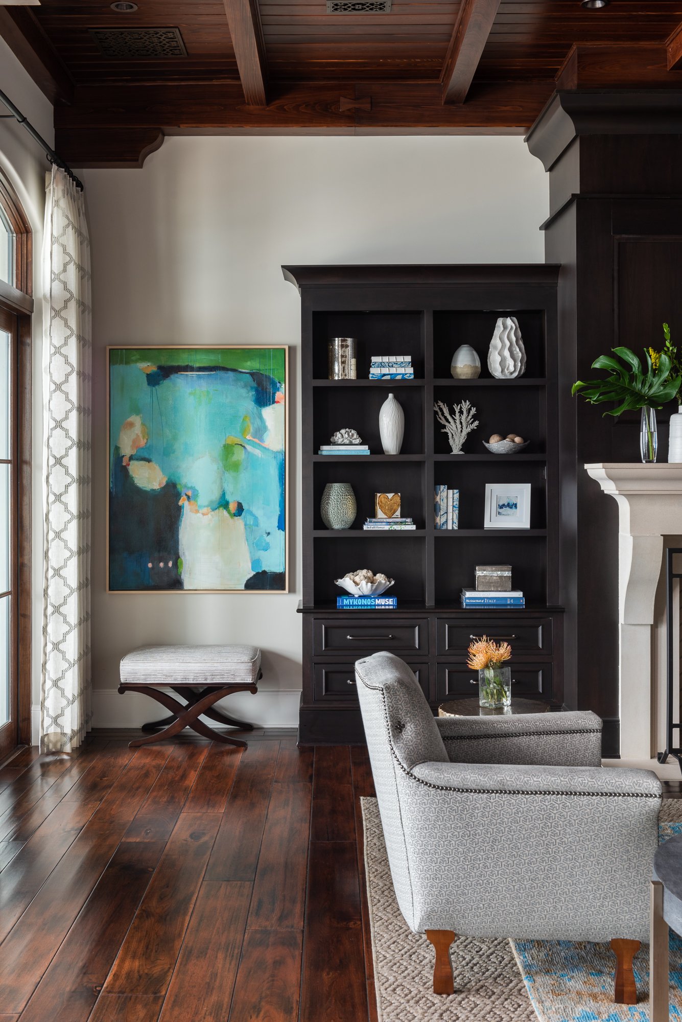 Interior of a living room with a dark wood bookshelf filled with decorative items and books, a gray armchair with a small coffee table holding a flower vase, a fireplace, an abstract painting, and wooden flooring.