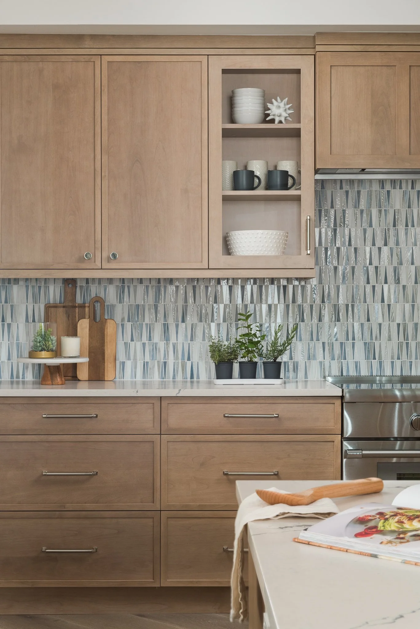 A modern kitchen with light wood cabinets, a tiled backsplash, and a marble countertop. Open shelves hold white and black cups and bowls. Potted plants and decorative items are on the counter. Part of a white kitchen island with a cookbook and a wood