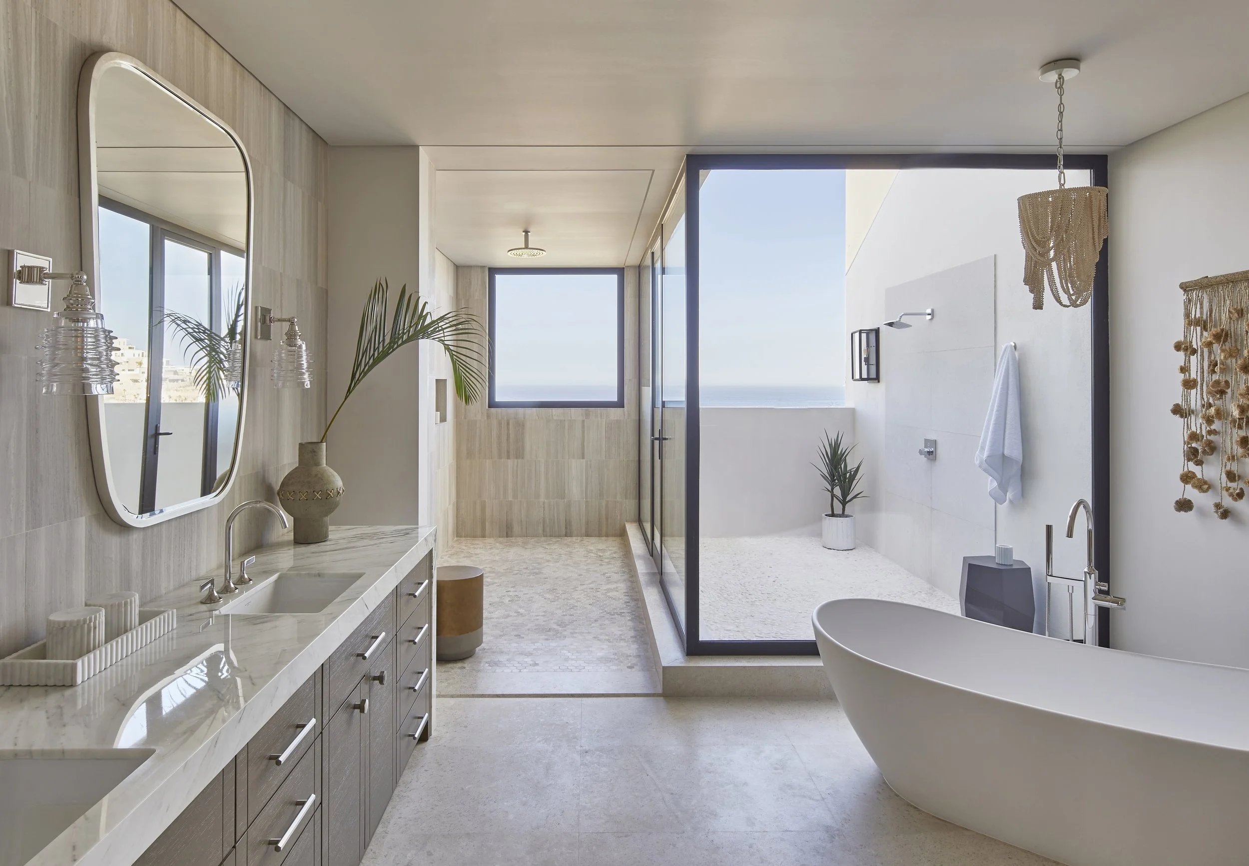 Modern bathroom with white freestanding bathtub, large walk-in shower with glass door, large window showing a sea view, beige tiled walls, and minimalistic decor including a vase with a plant, a mirror, and a small side table.