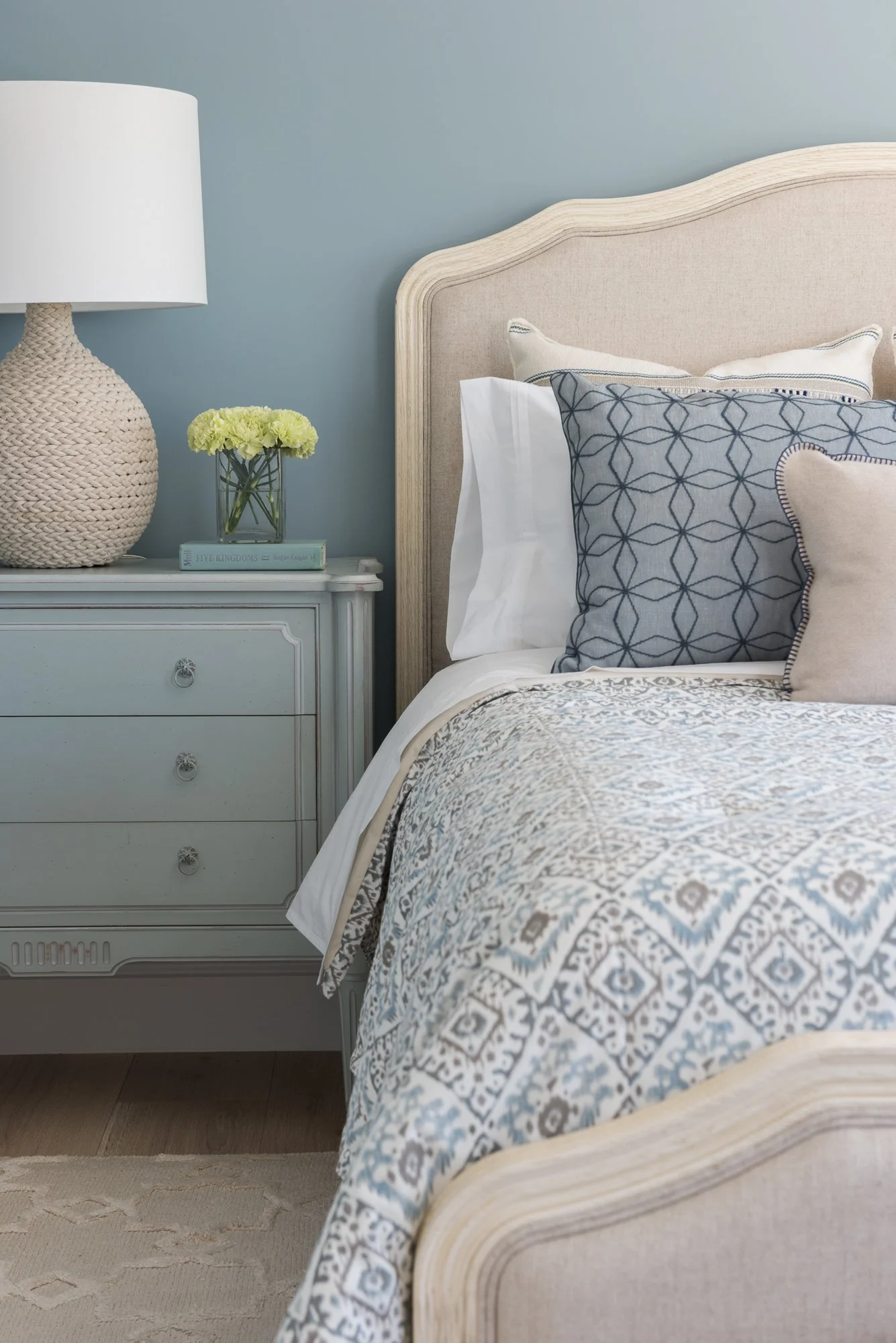 Close-up of a neatly made bed with patterned bedding and pillows, next to a light blue nightstand holding a lamp with a woven base and a small vase of green flowers.