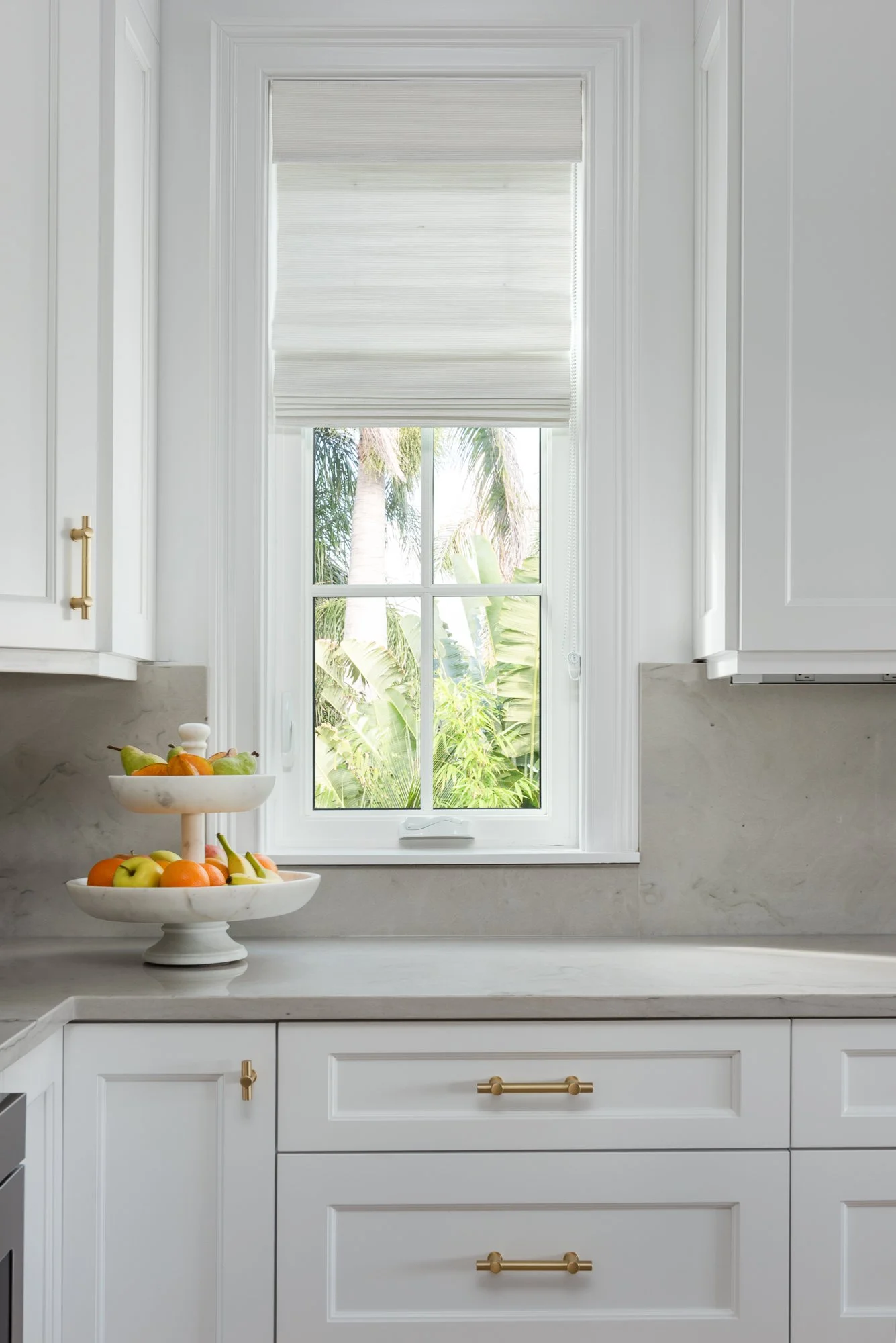 A bright kitchen countertop with a tiered fruit stand and a window showing palm trees outside.