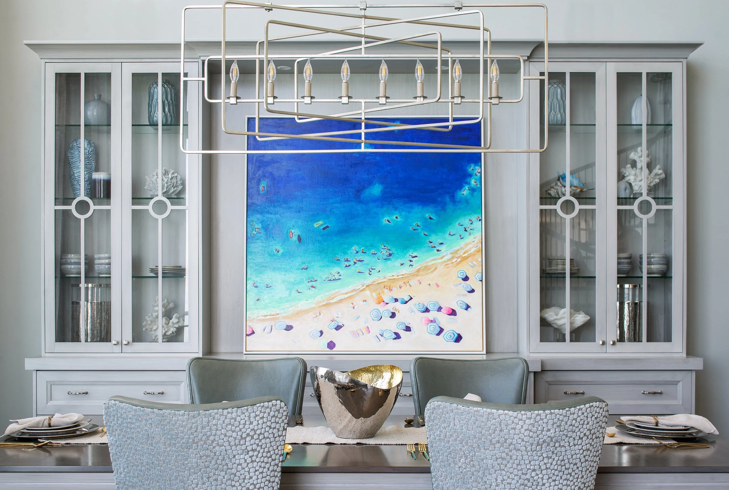A dining room with a large abstract beach scene painting on a white shelf with glass doors. The shelf contains decorative items such as vases, coral, and dishes. There is a modern chandelier hanging above the table with a metallic bowl in the center 