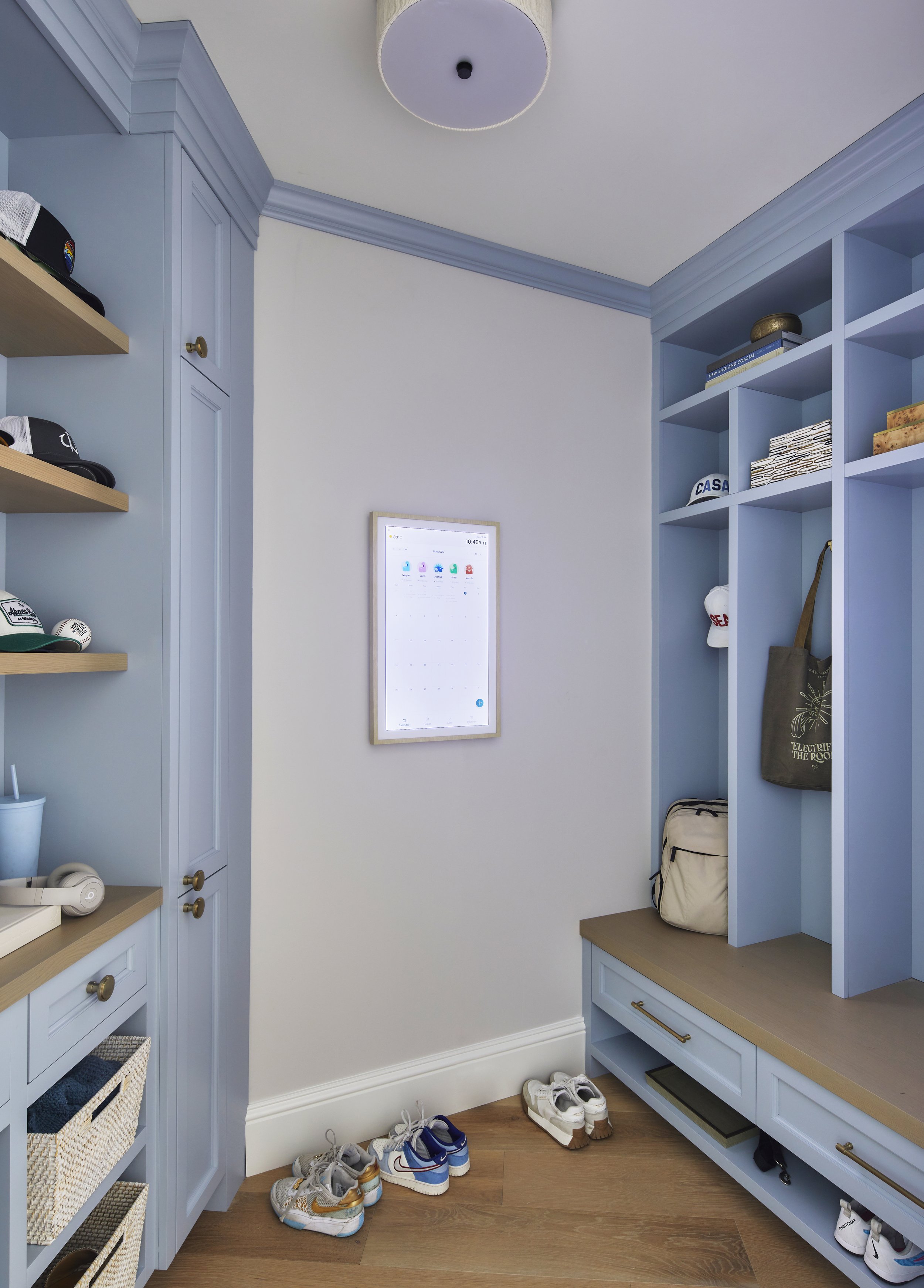 Entryway with light blue built-in shelves and cabinets, a white wall with a framed digital calendar, shoes on the wooden floor, and a ceiling light fixture.