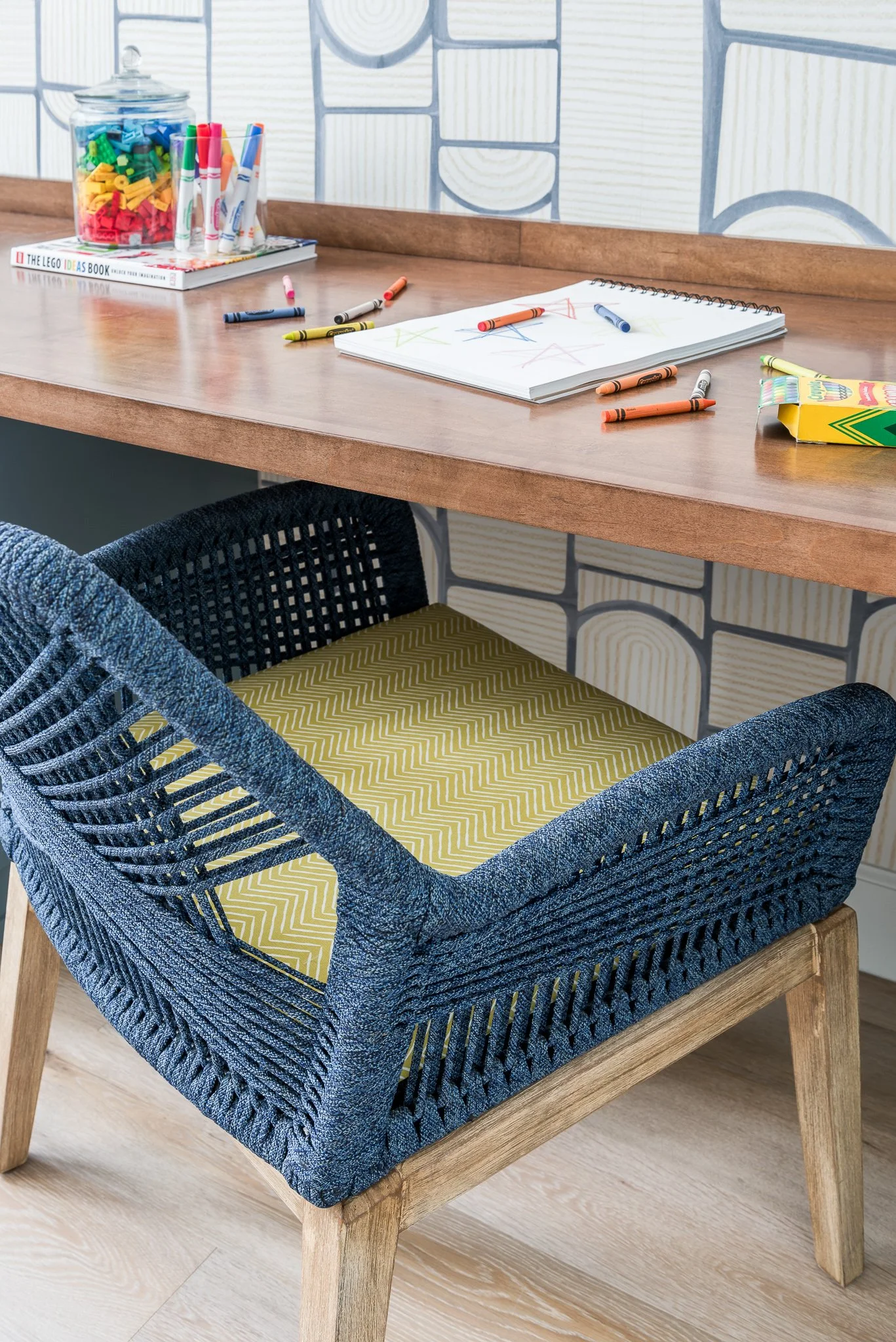 A wooden desk with scattered crayons, a notebook with crayon drawings, a jar of colorful building blocks, and a box of crayons, with a patterned wallpaper in the background and a woven chair with a yellow cushion underneath.