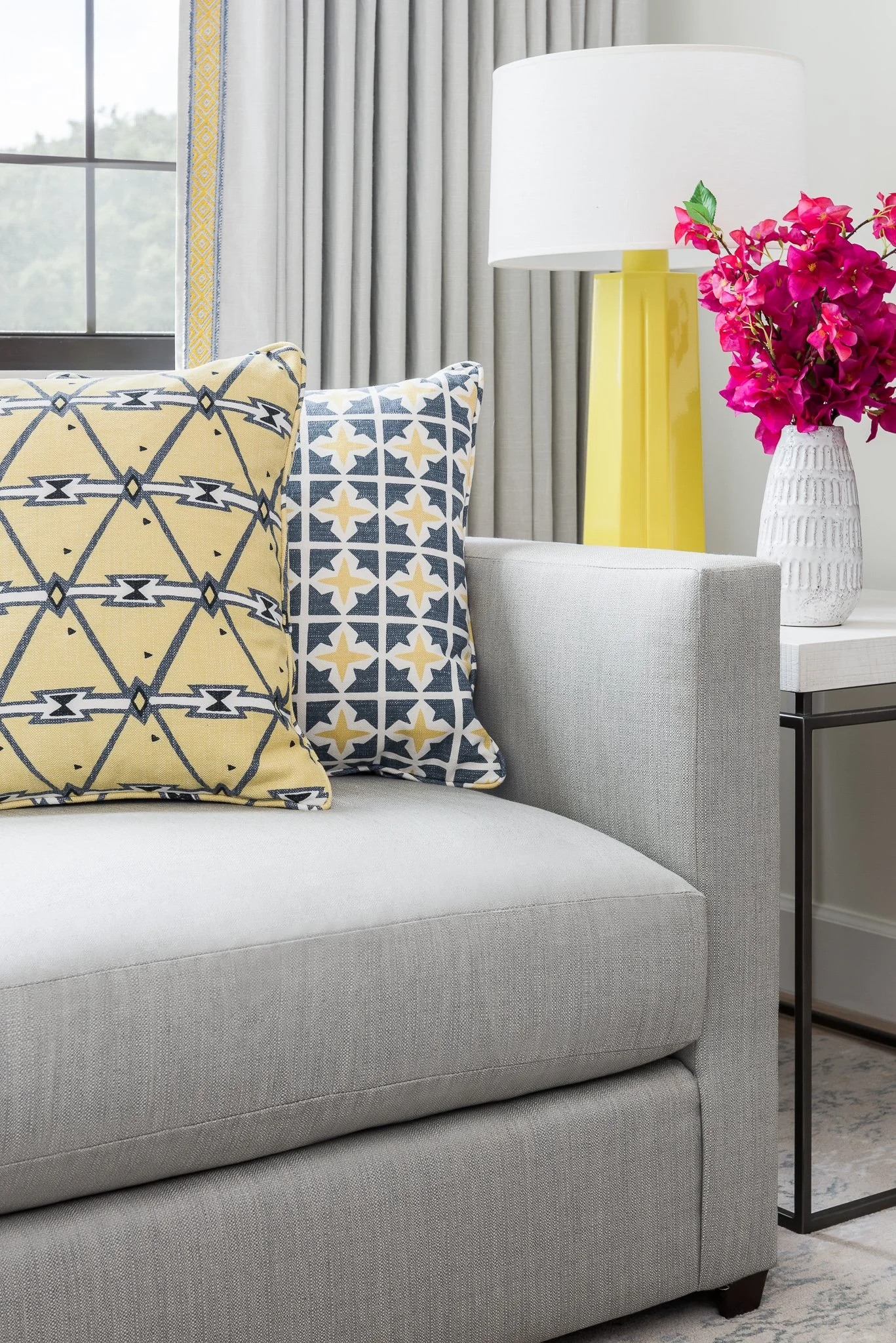 Close-up of a modern living room corner with decorative pillows on a light gray sofa, a yellow lamp, pink flowers in a white vase, and curtains in the background.