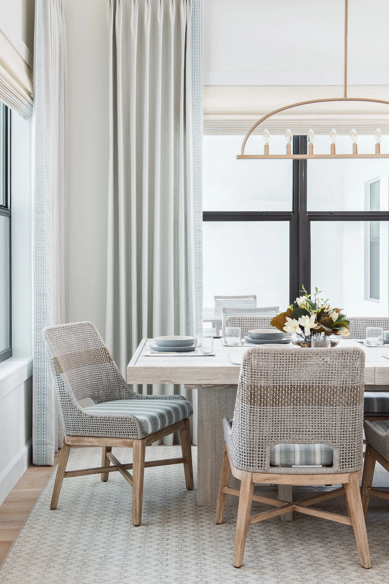 Dining area with a wooden table, woven chairs with striped cushions, a flower centerpiece, and a window with curtains and blinds
