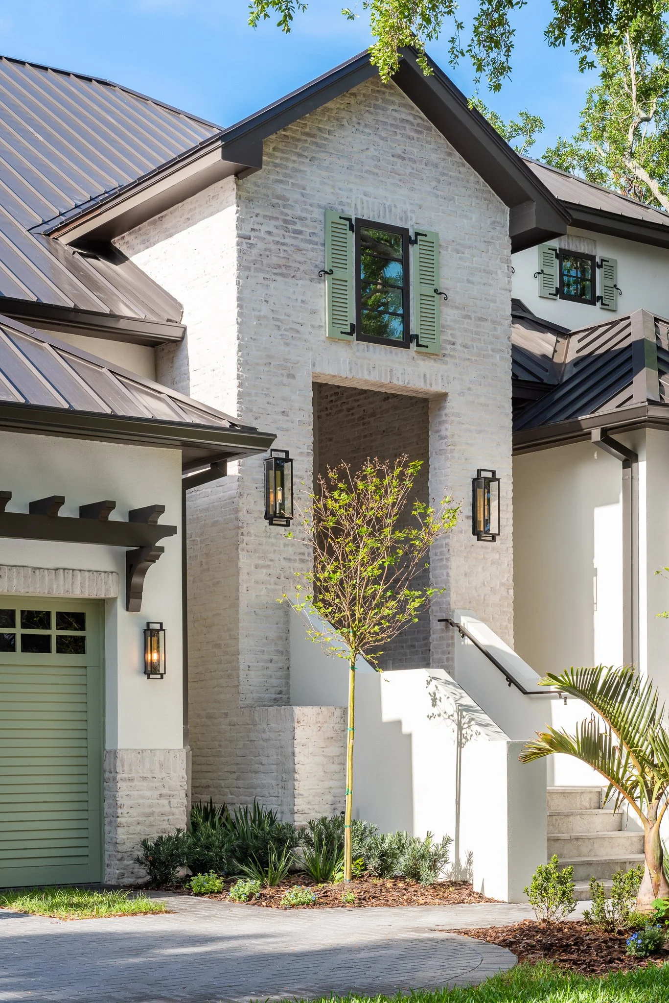 Exterior of a modern house with a brick and stucco facade, black-framed windows with shutters, a staircase with a black railing, and landscaped garden with a small tree, plants, and green grass.