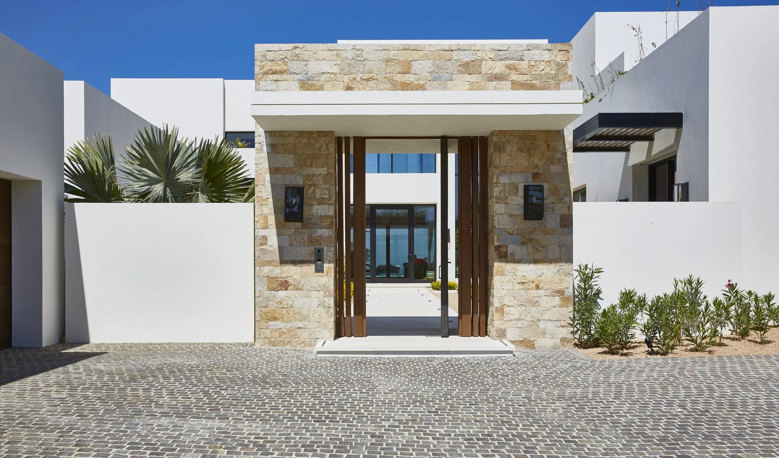 Modern house entrance made of stone and white walls with wooden gate and potted plants, sunny day with blue sky.