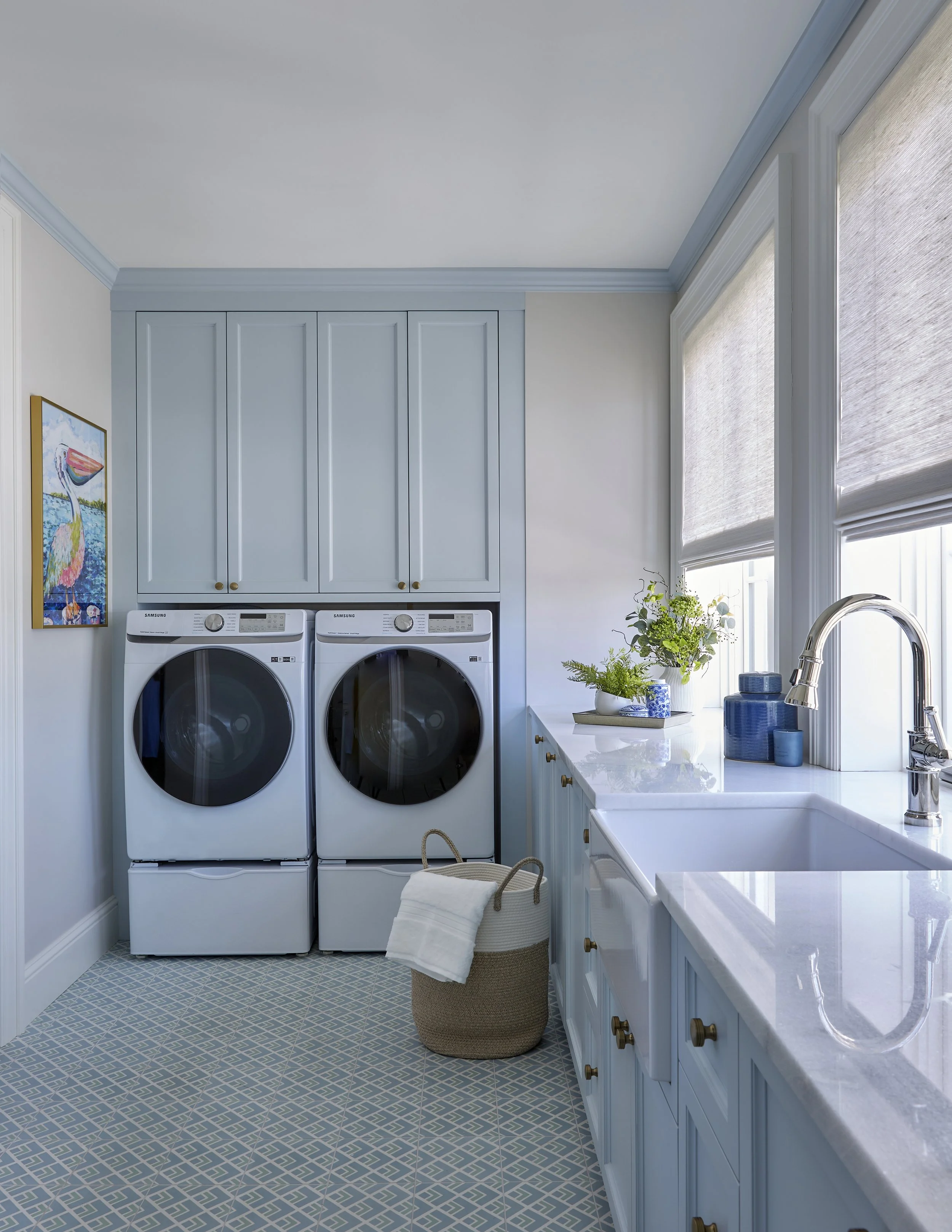 A laundry room with a white countertop, blue cabinets, a sink, and a window with beige shades. Two Samsung washing machines are positioned side by side with a hanging laundry basket on the floor in front of them. There are plants and decorative items