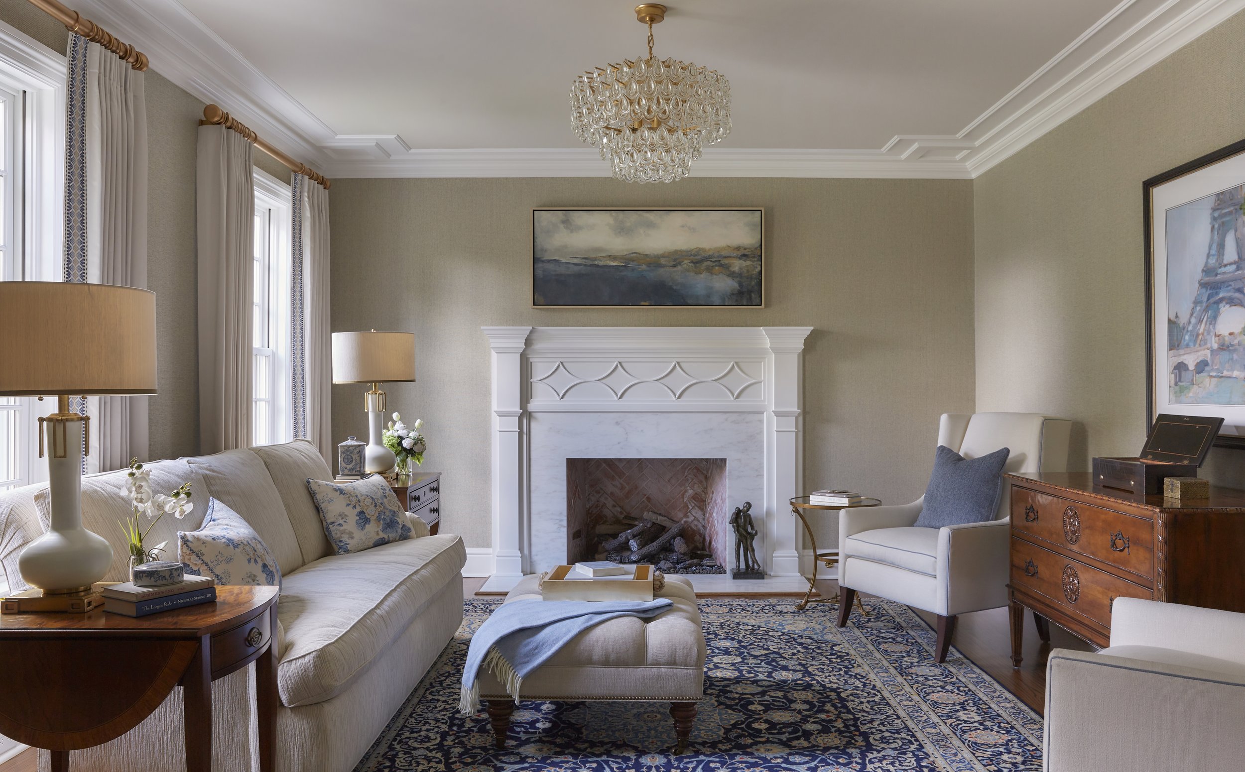 Elegant living room with cream-colored sofa, white armchair, and fireplace with a painting above. There are lamps, a wooden side table, and decorative pillows, along with a patterned rug on the wooden floor.