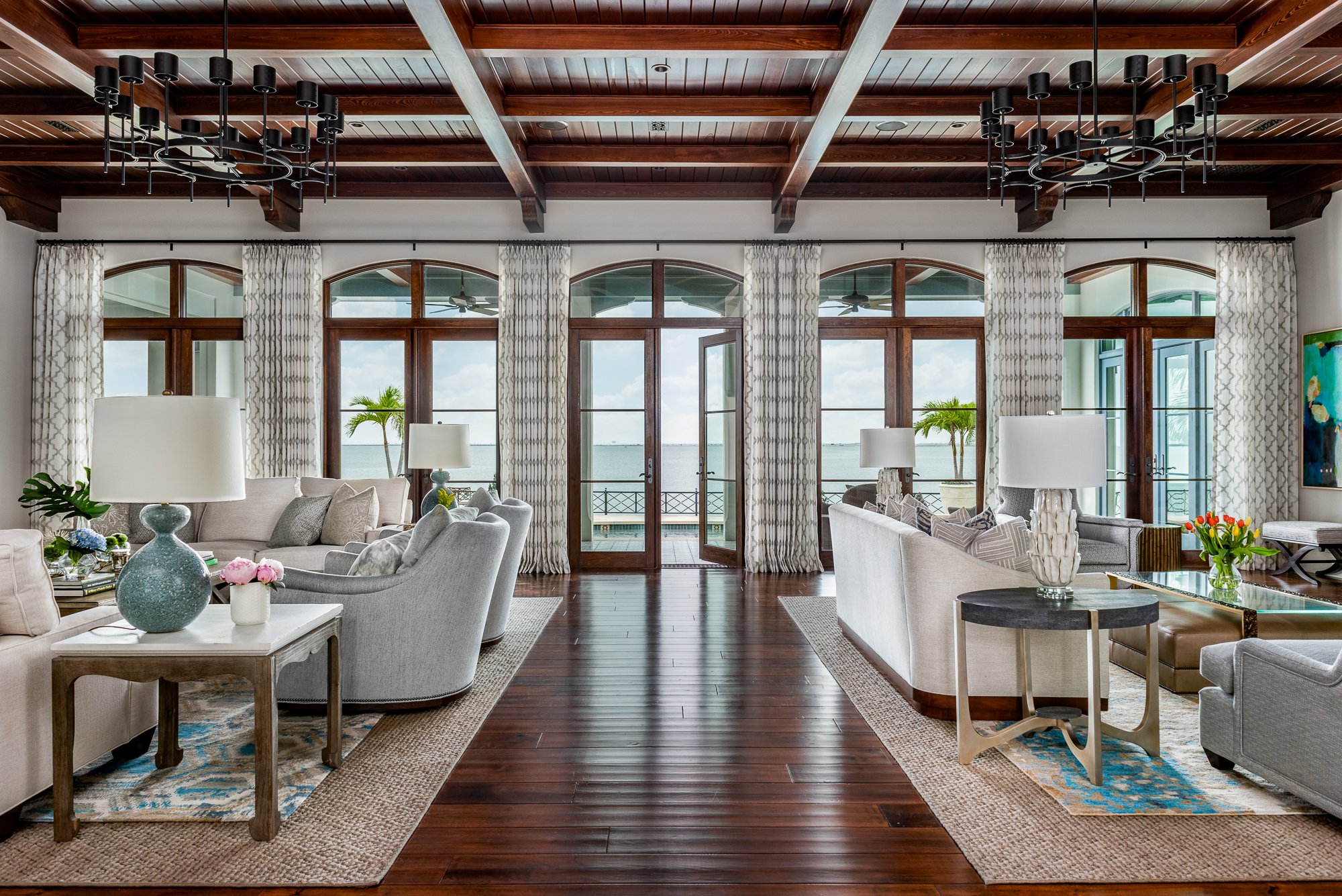 Living room with large windows overlooking water, wooden flooring, modern chandeliers, and comfortable sofas.