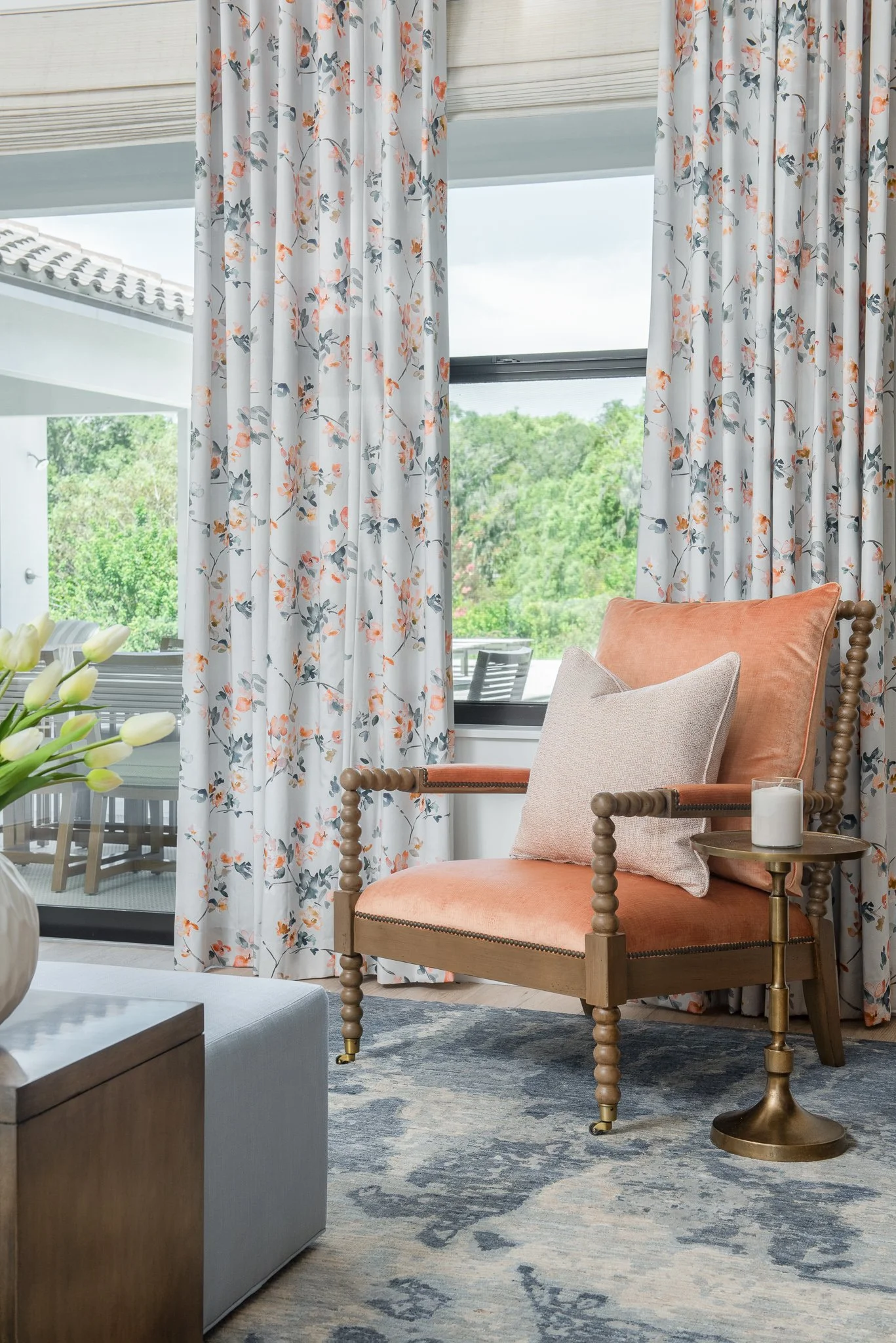 A cozy living room corner with a peach-colored armchair, beige pillow, small side table holding a candle, floral curtains, and large windows showing greenery outside.