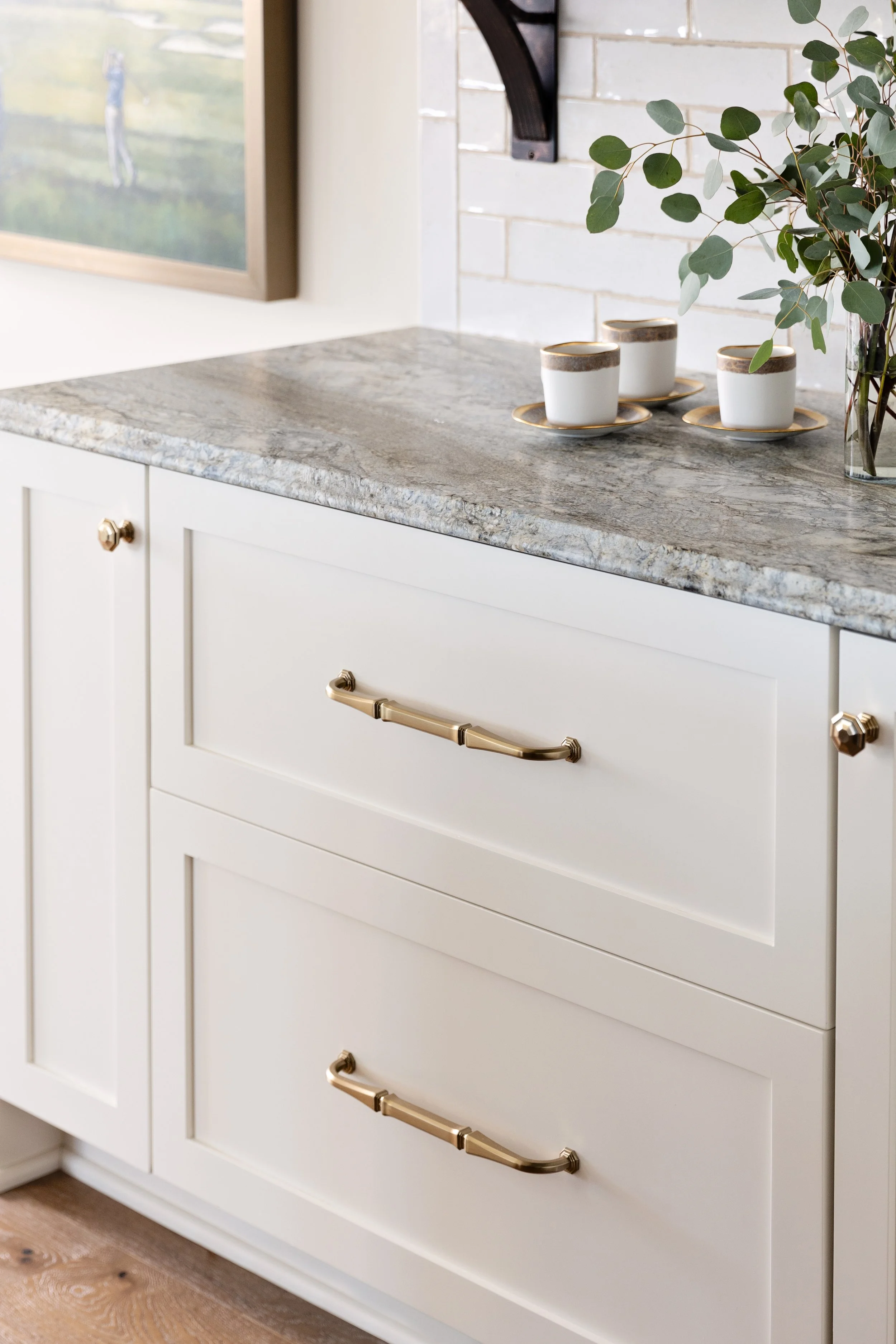White kitchen cabinet with granite countertop, three golden handles, a plant in a glass vase, and three white coffee cups with gold rims on saucers.