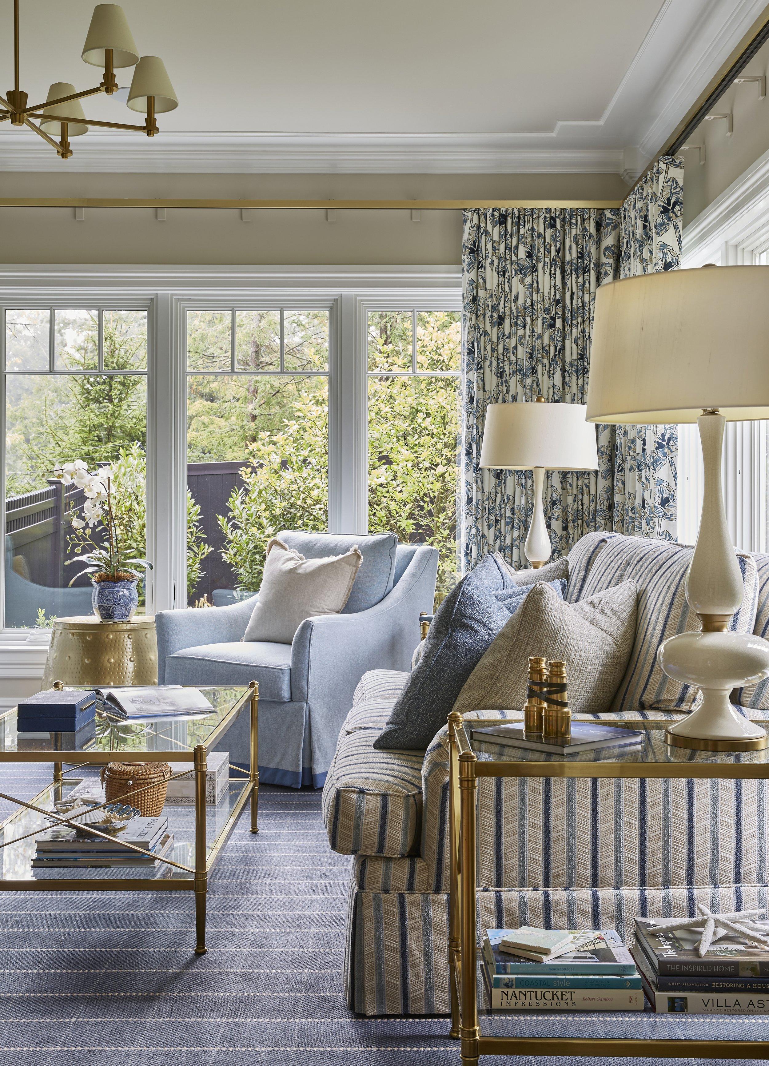 Living room with large windows, striped sofa, blue armchair, gold side tables, table lamps, and books.