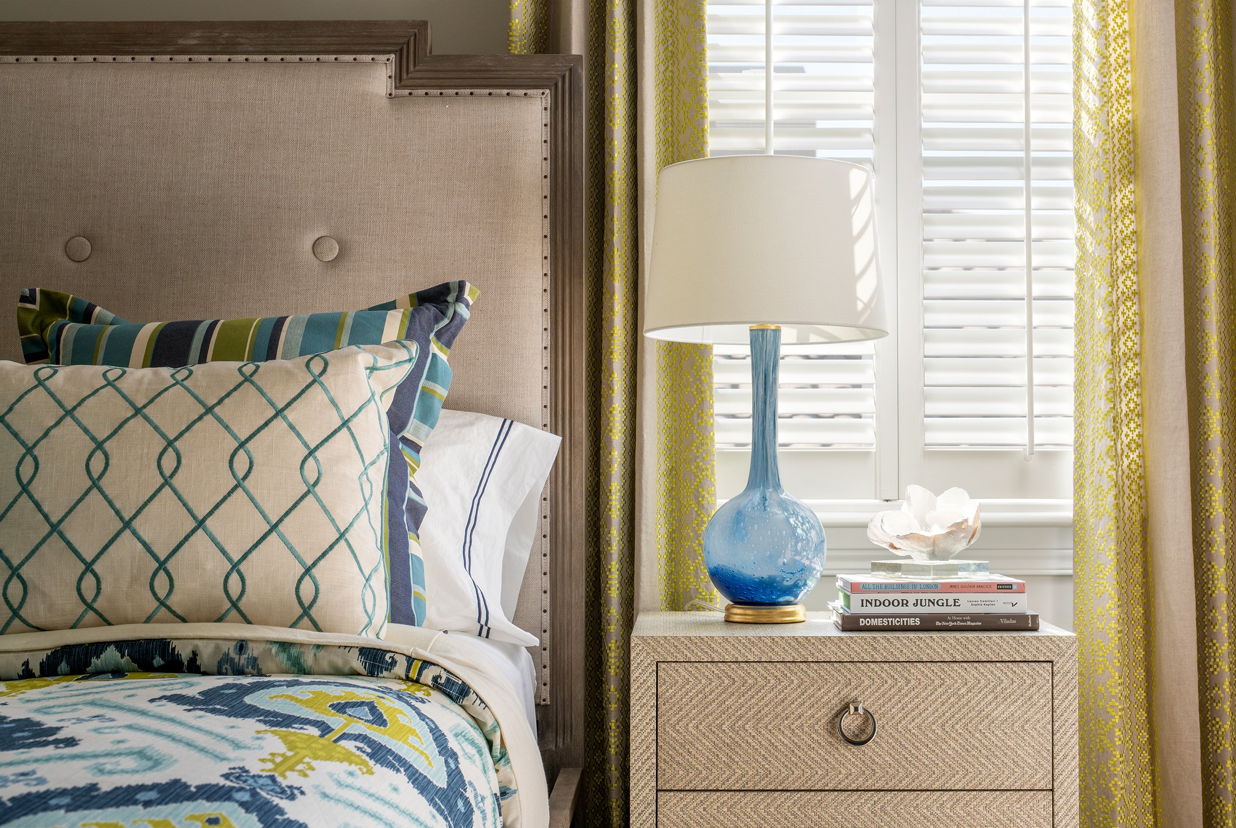 A bedroom with a beige upholstered headboard, colorful pillows with blue, green, and white patterns, and a patterned bedspread. A nightstand with a blue glass lamp, a stack of books, and a decorative shell sculpture sits by a window with white shutte