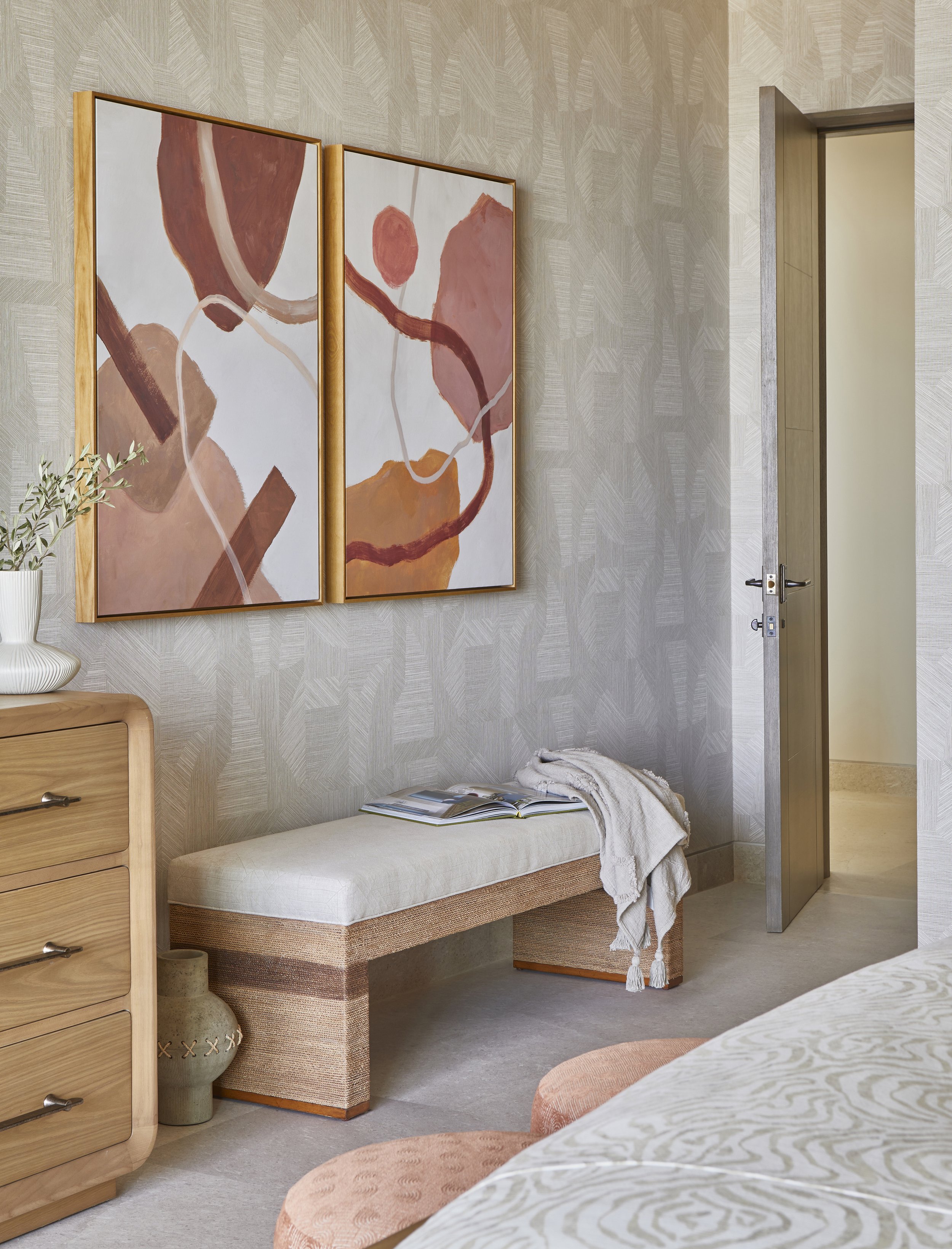 Interior of a bedroom with abstract wall art, a wooden dresser with a plant, a beige bench with an open book and a scarf, and a partially visible bed with patterned bedding.