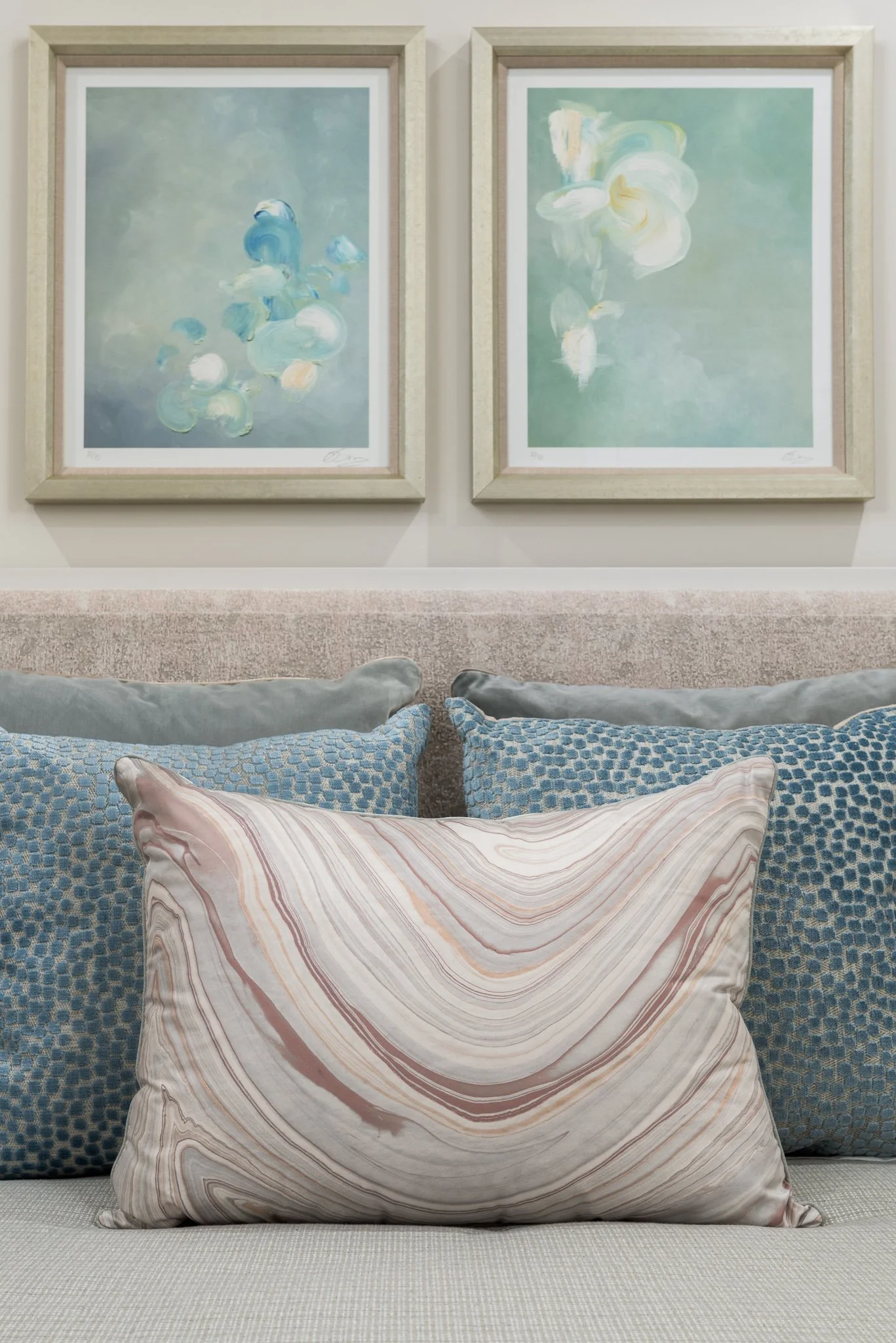 A beige sofa with four decorative pillows in front of a beige wall with two framed abstract paintings. The pillows are pinkish marble pattern, blue with a checkered pattern, and plain grey.