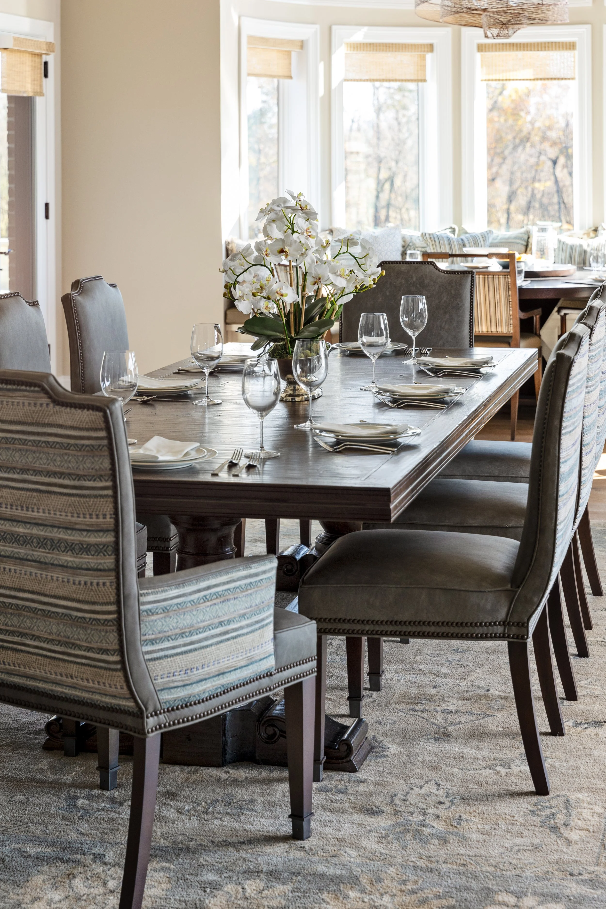 Elegant dining room with a wooden table set for six, featuring white plates, silverware, wine glasses, and a large floral centerpiece of white orchids, with a sunlit window in the background.