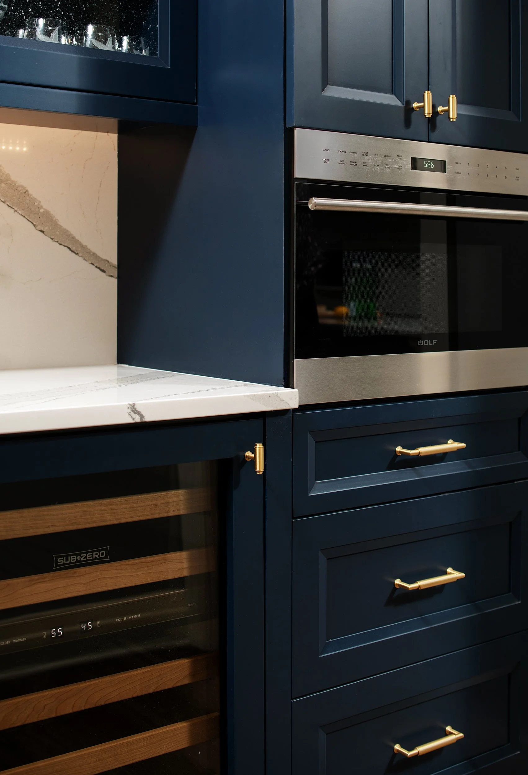 Close-up of modern kitchen cabinets painted dark blue with gold handles, a built-in microwave, marble countertop, and a wine fridge with wood accents.