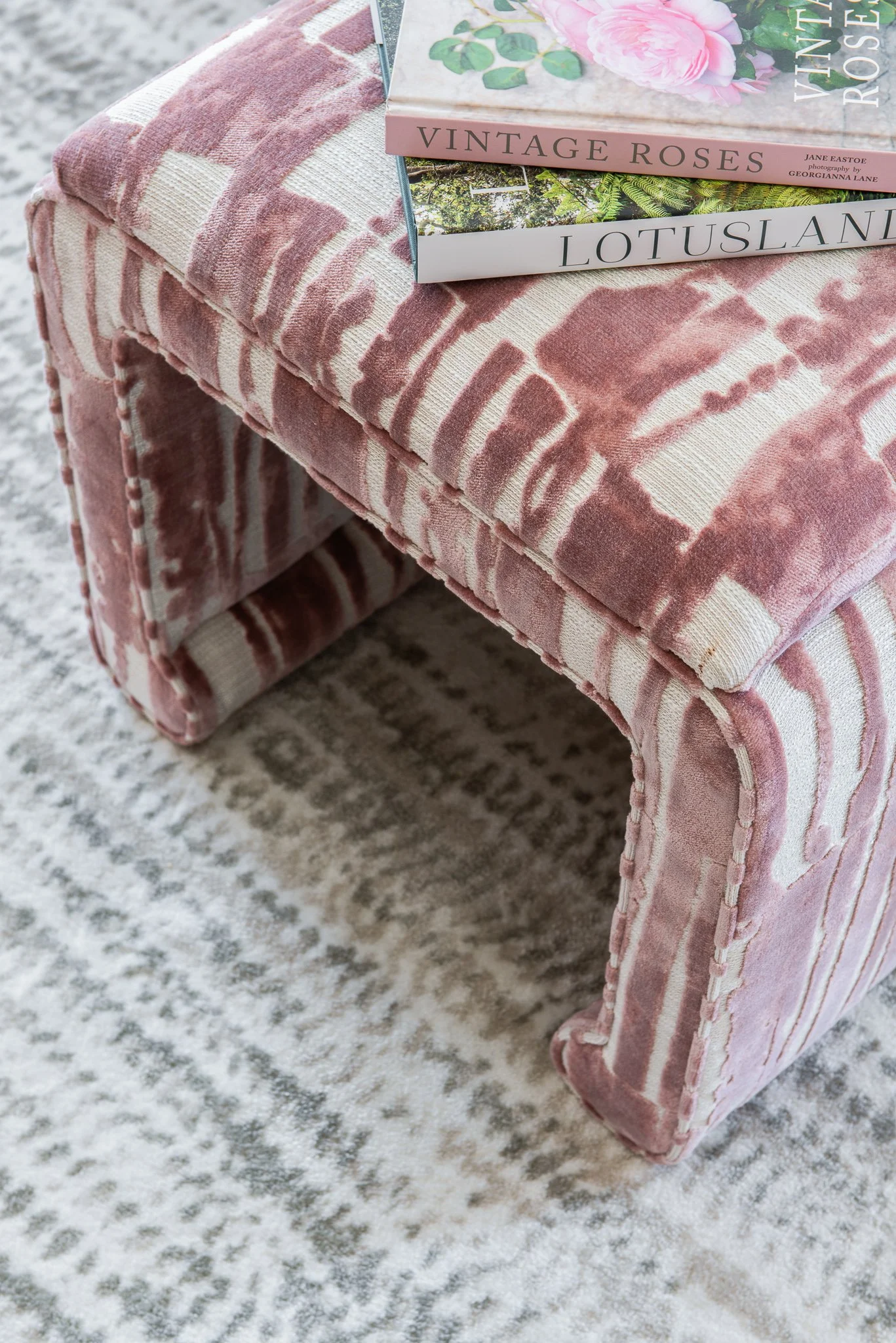 Close-up of a pink patterned upholstered bench with two decorative hardcover books on top, placed on a light patterned rug.
