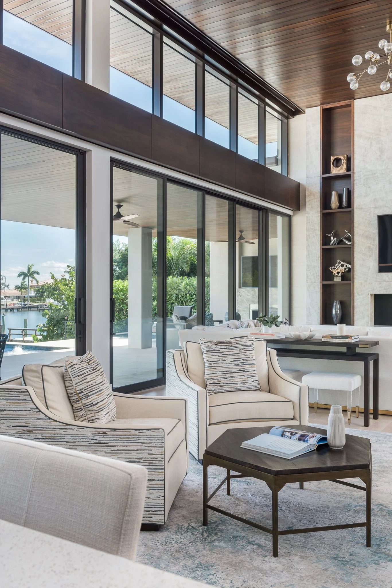 Modern living room with large glass sliding doors, beige sofas with patterned pillows, a dark wood coffee table with a book and a white vase, and a built-in wall shelf with decorative items. Outdoor view of greenery and water is visible.