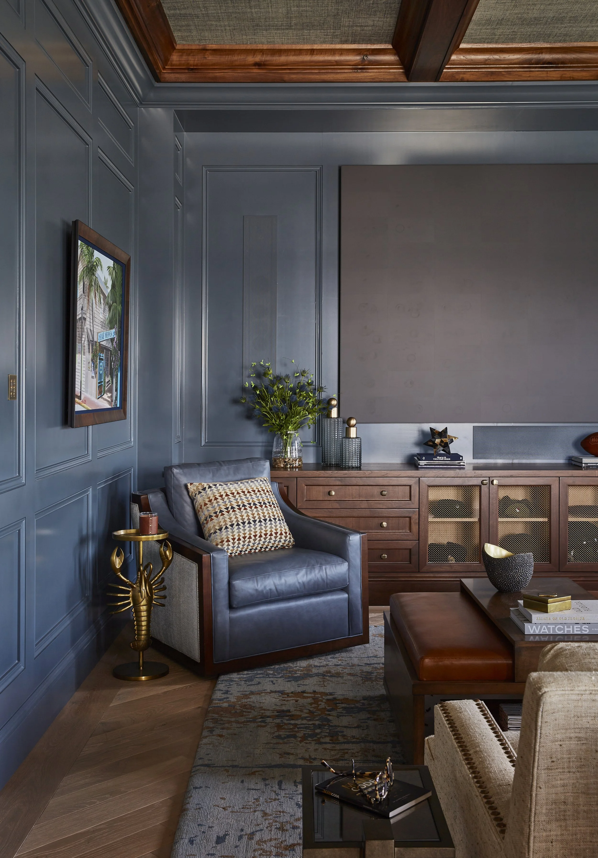 Living room with blue paneled walls, wooden ceiling, and hardwood floor. Contains a black leather armchair with a patterned pillow, a wooden side table with a gold lobster-shaped base and a candle, a wooden cabinet with decorative vases and books, a 