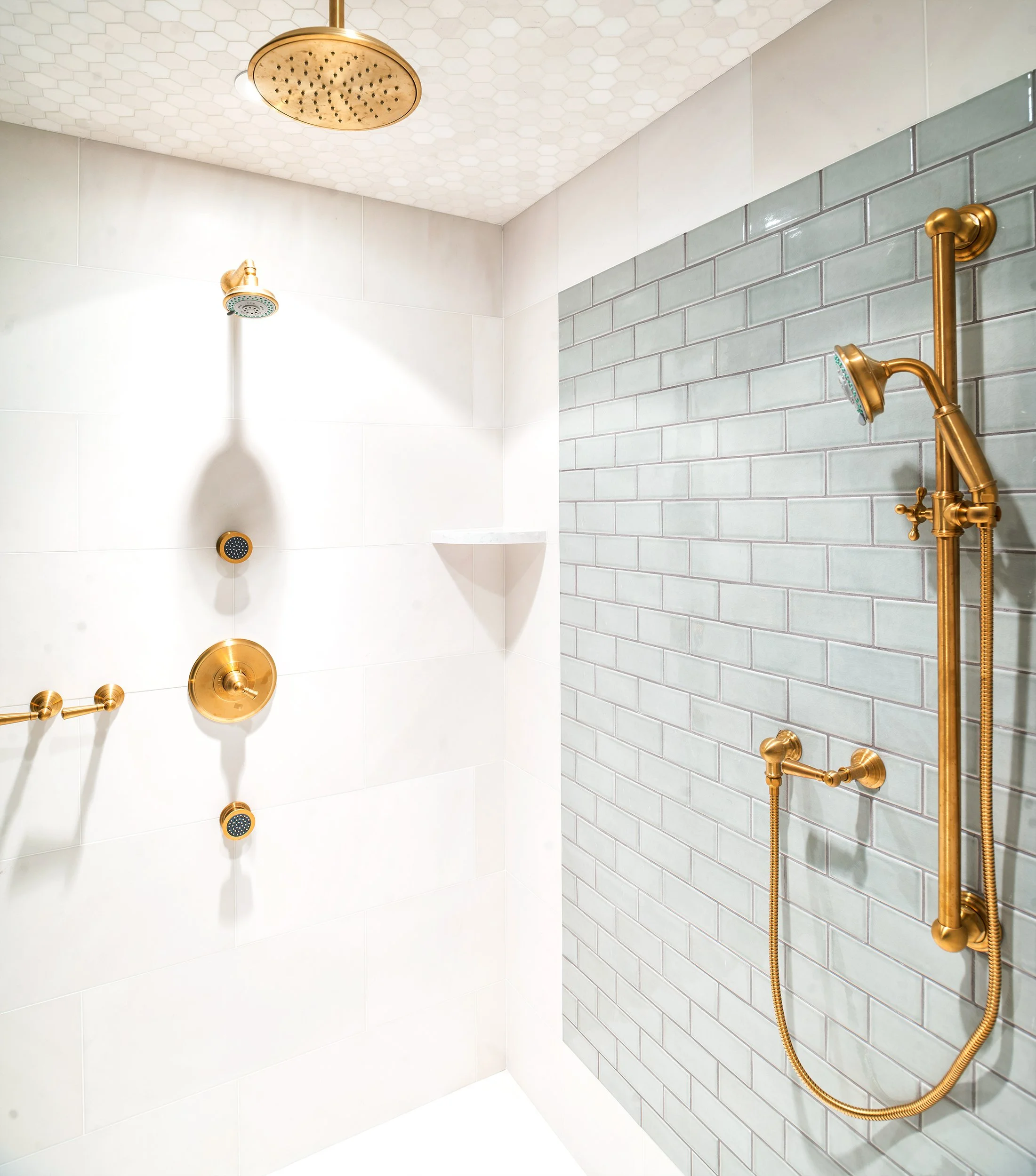 Modern shower with white tiles, a gray brick accent wall, and gold fixtures including a showerhead, handheld shower, and controls.