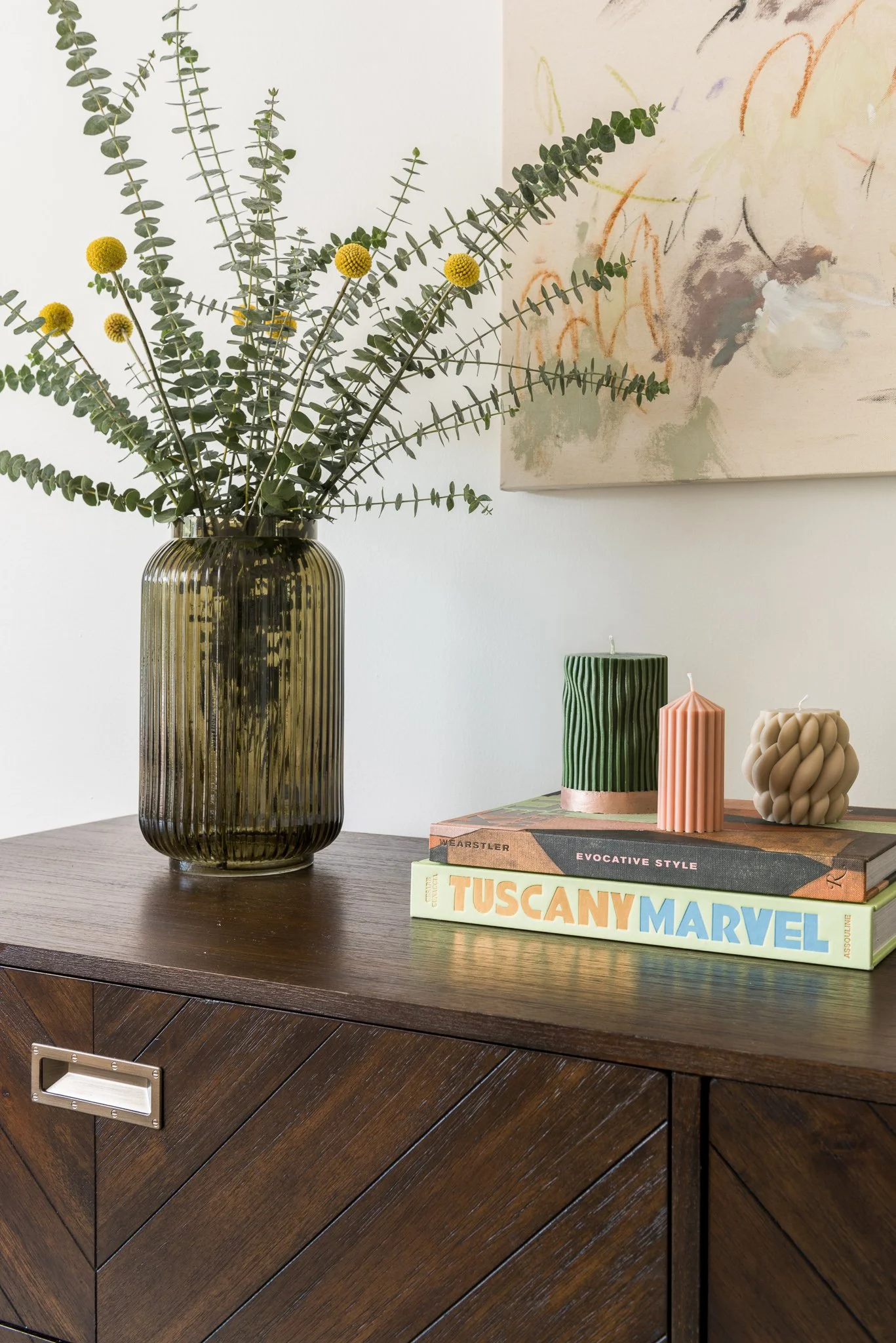 Decorative arrangement on a wooden sideboard featuring a large ribbed glass vase filled with greenery and yellow flowers, three candles in different shapes and colors, and colorful books.