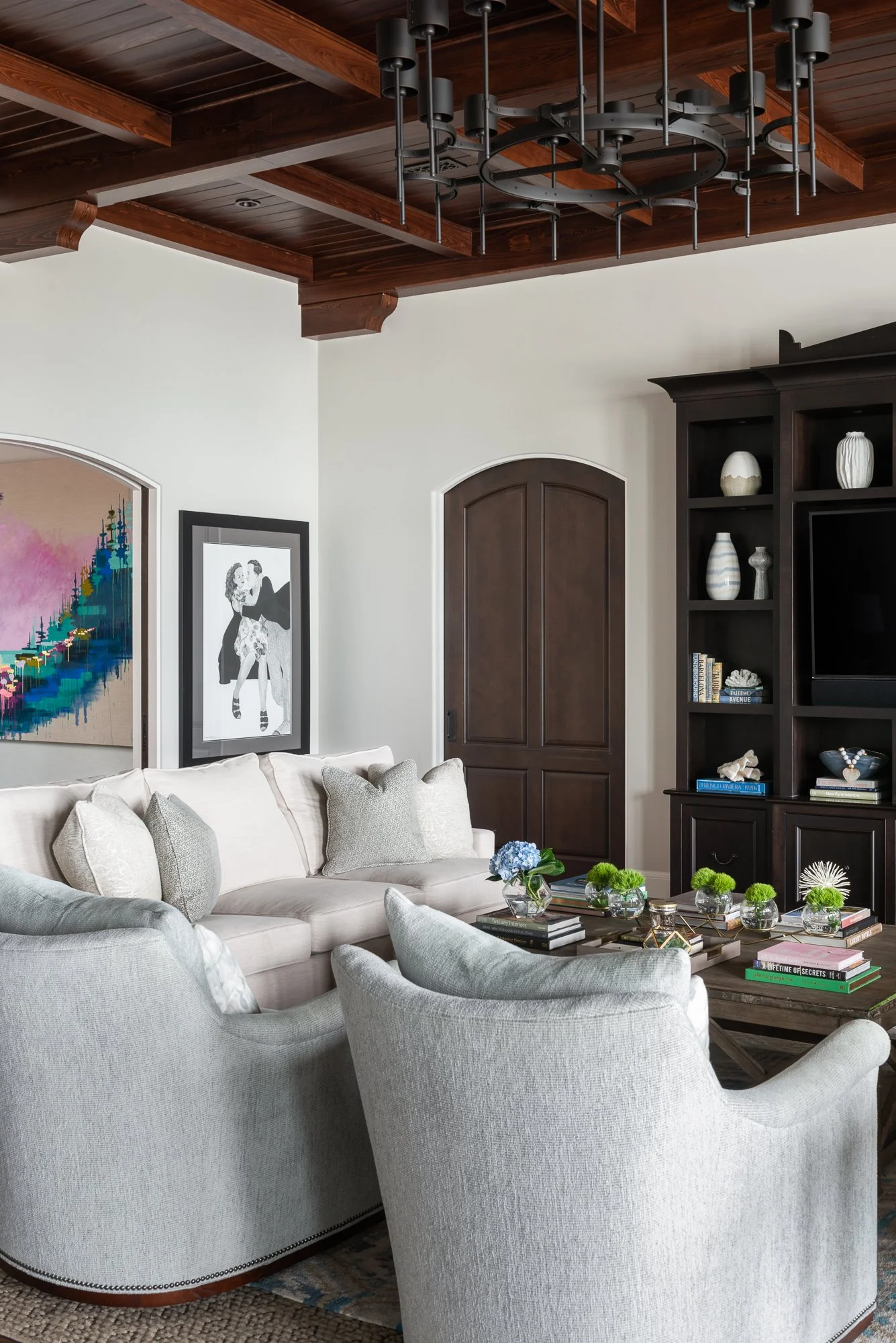 Living room with white sofa and armchair, dark wood coffee table with decorative items and books, built-in dark wood shelving, white walls with art, and a wooden ceiling with black metal chandelier.