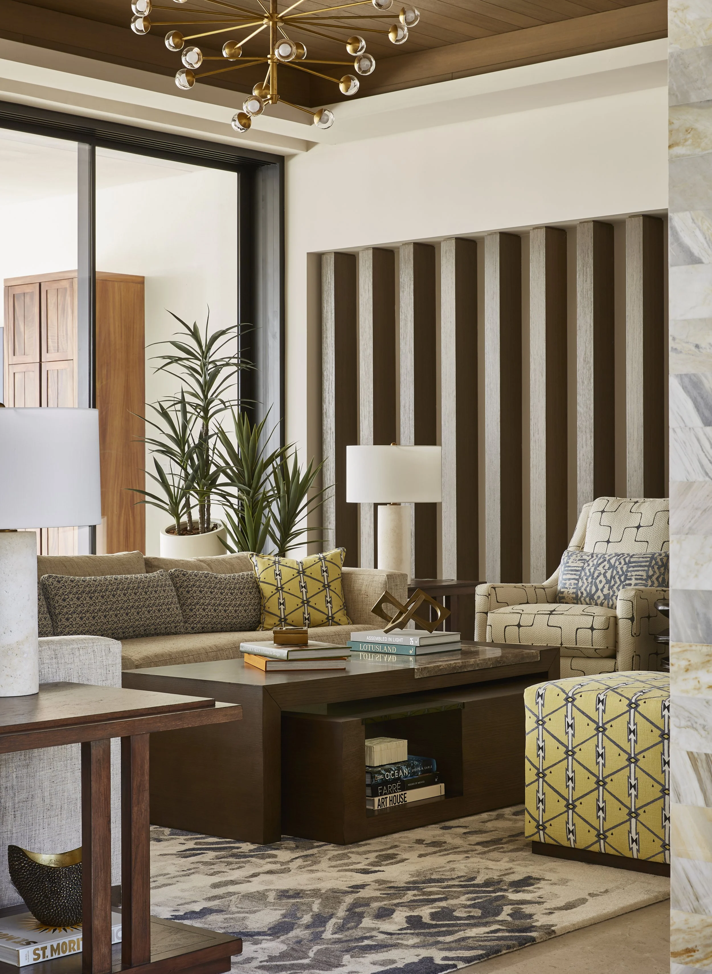 Living room with beige sofa, patterned armchair, wooden coffee table, table lamp, tall potted plants, rug, and modern chandelier.