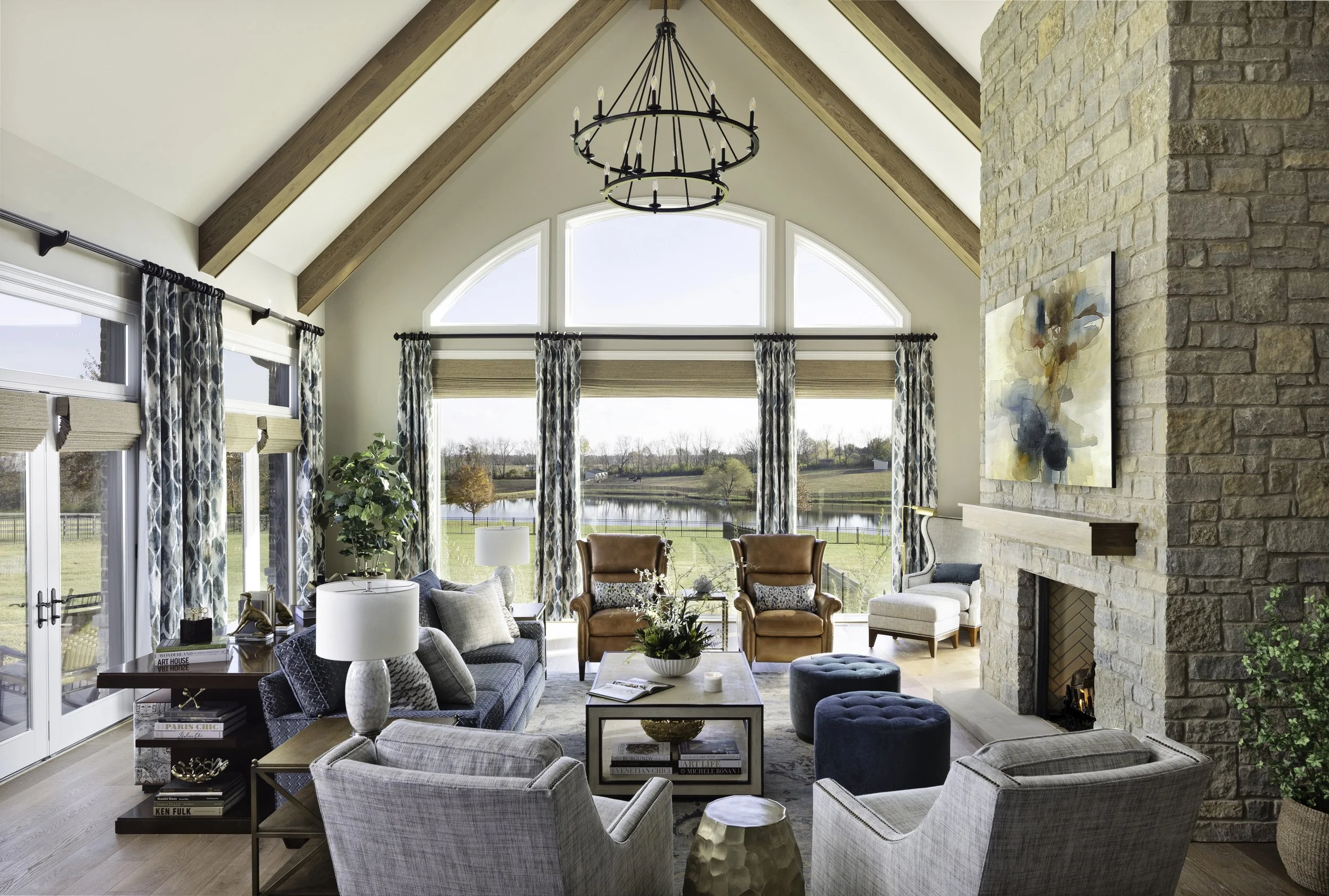 Living room with large arched windows overlooking a lake, featuring a stone fireplace, chandelier, and various chairs and sofas.