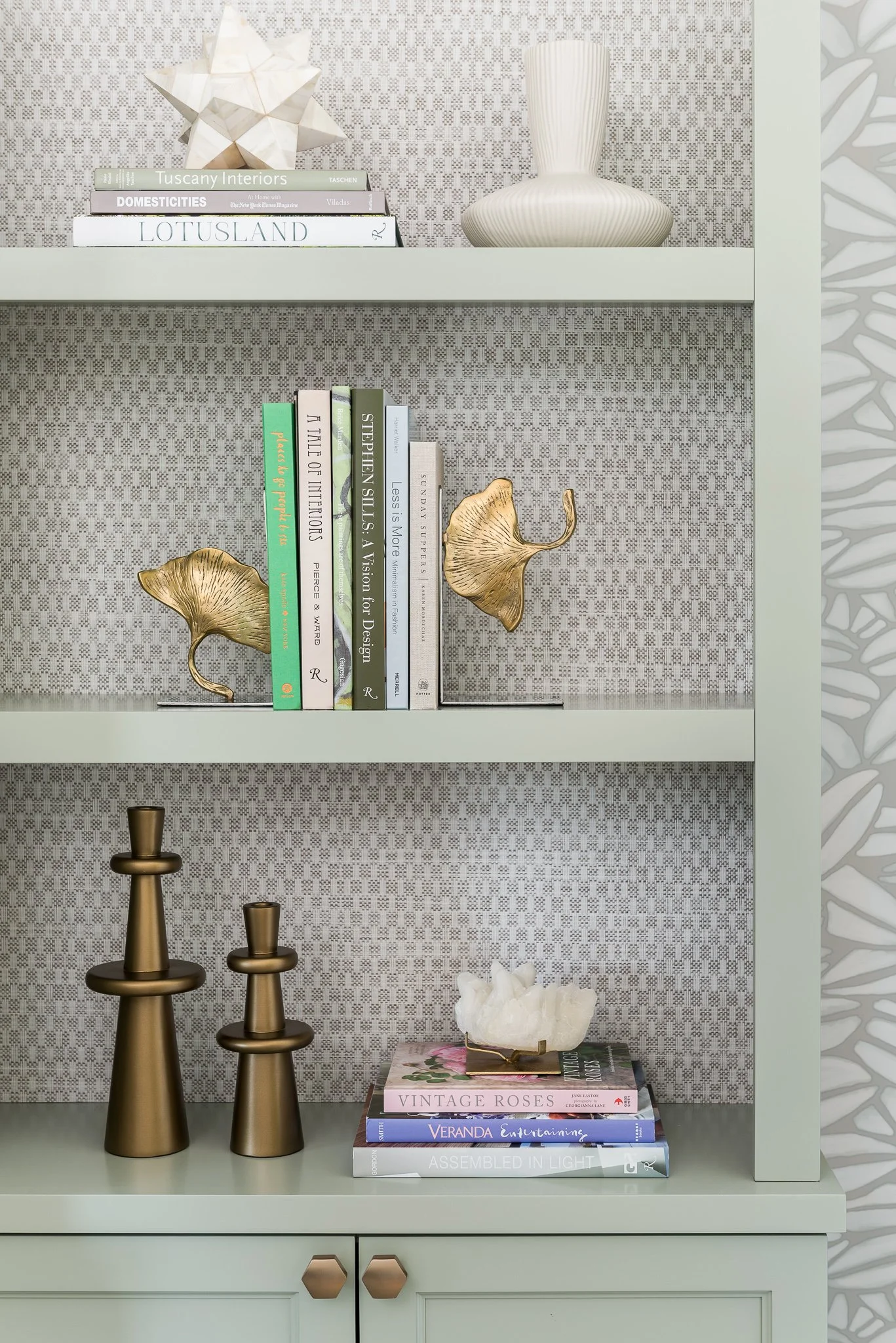 A light green bookshelf with three shelves, decorated with books, gold and white decorative objects, and a patterned background.