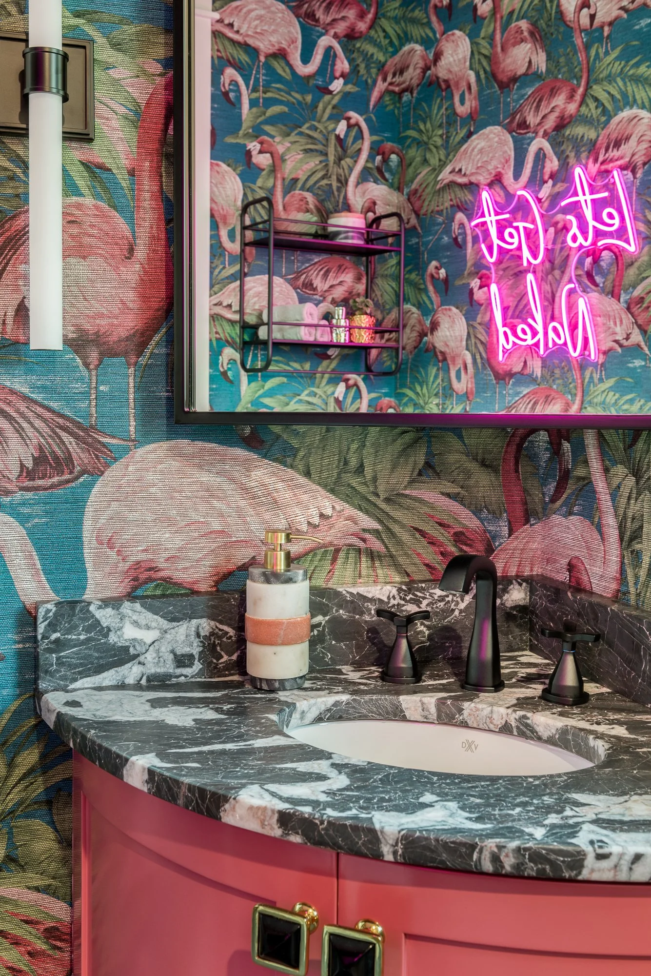 A bathroom with tropical flamingo wallpaper, a pink neon sign, a black marble countertop with an oval sink, a soap dispenser, and pink cabinets with gold handles.
