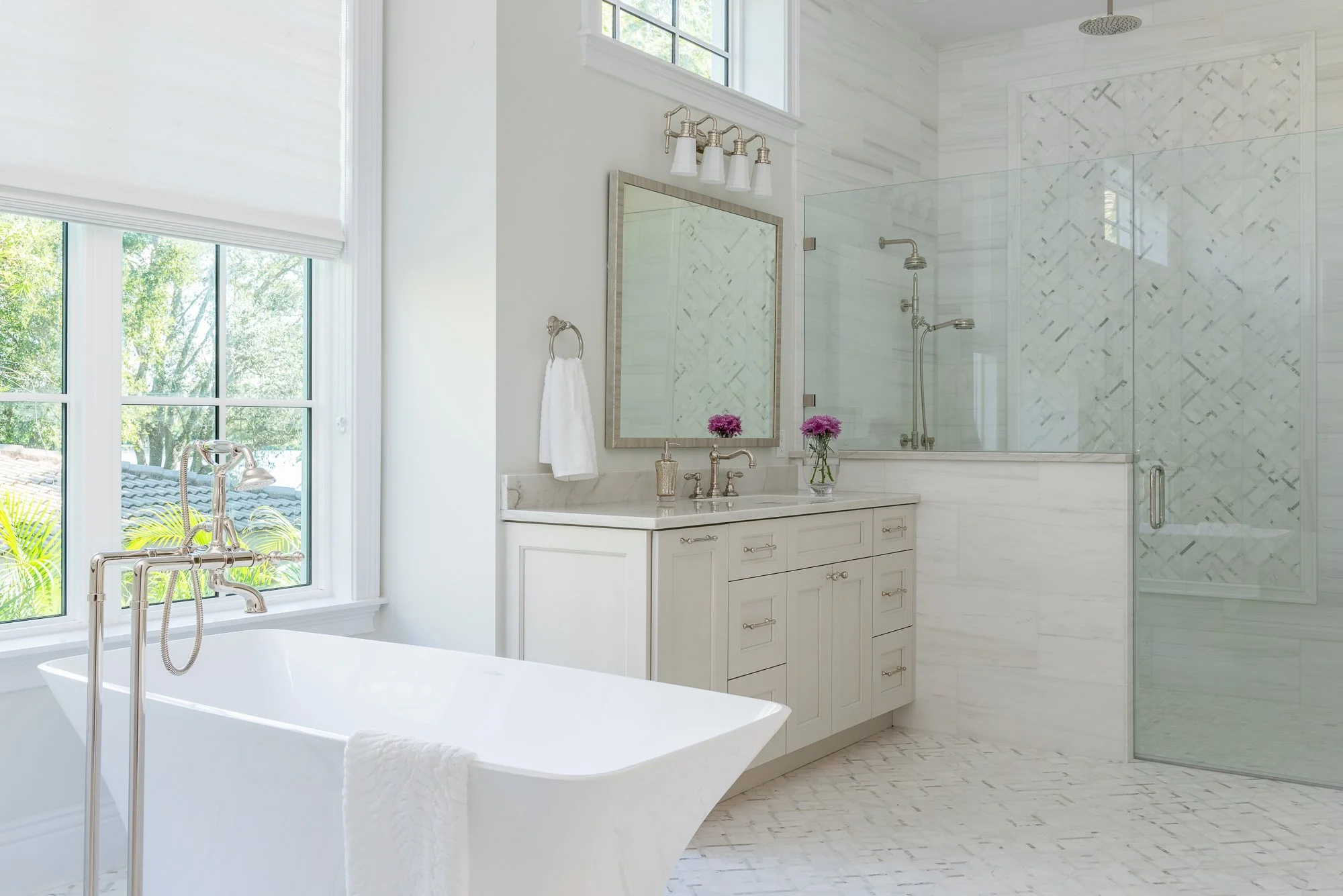 Bright bathroom with a white freestanding bathtub near a large window, a vanity with a mirror and sink, and a walk-in shower with glass door and tiled walls.