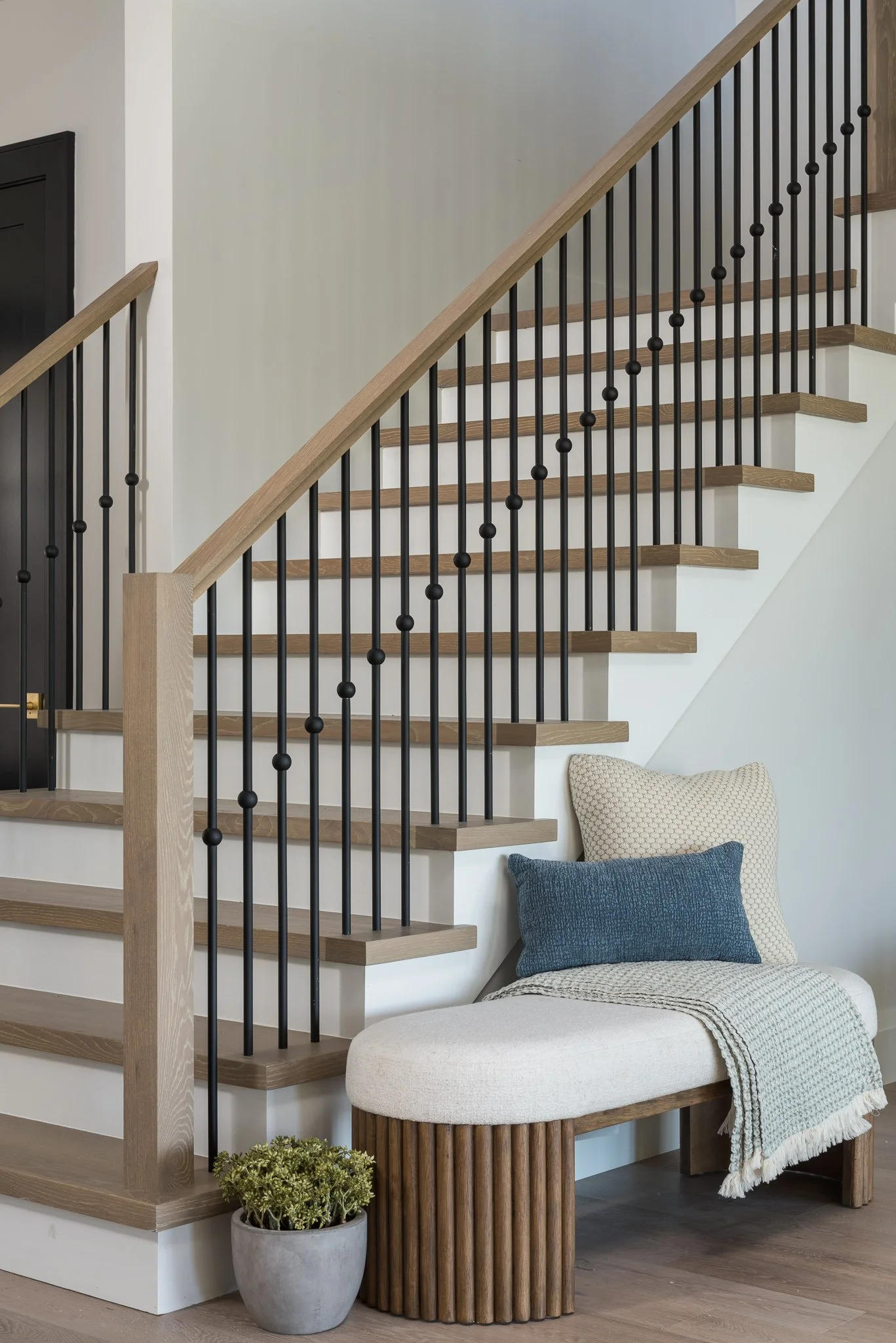 Interior view of a modern staircase with wooden steps and black metal railing, next to a cozy seating nook with a curved bench, pillows, and a plant in a pot.